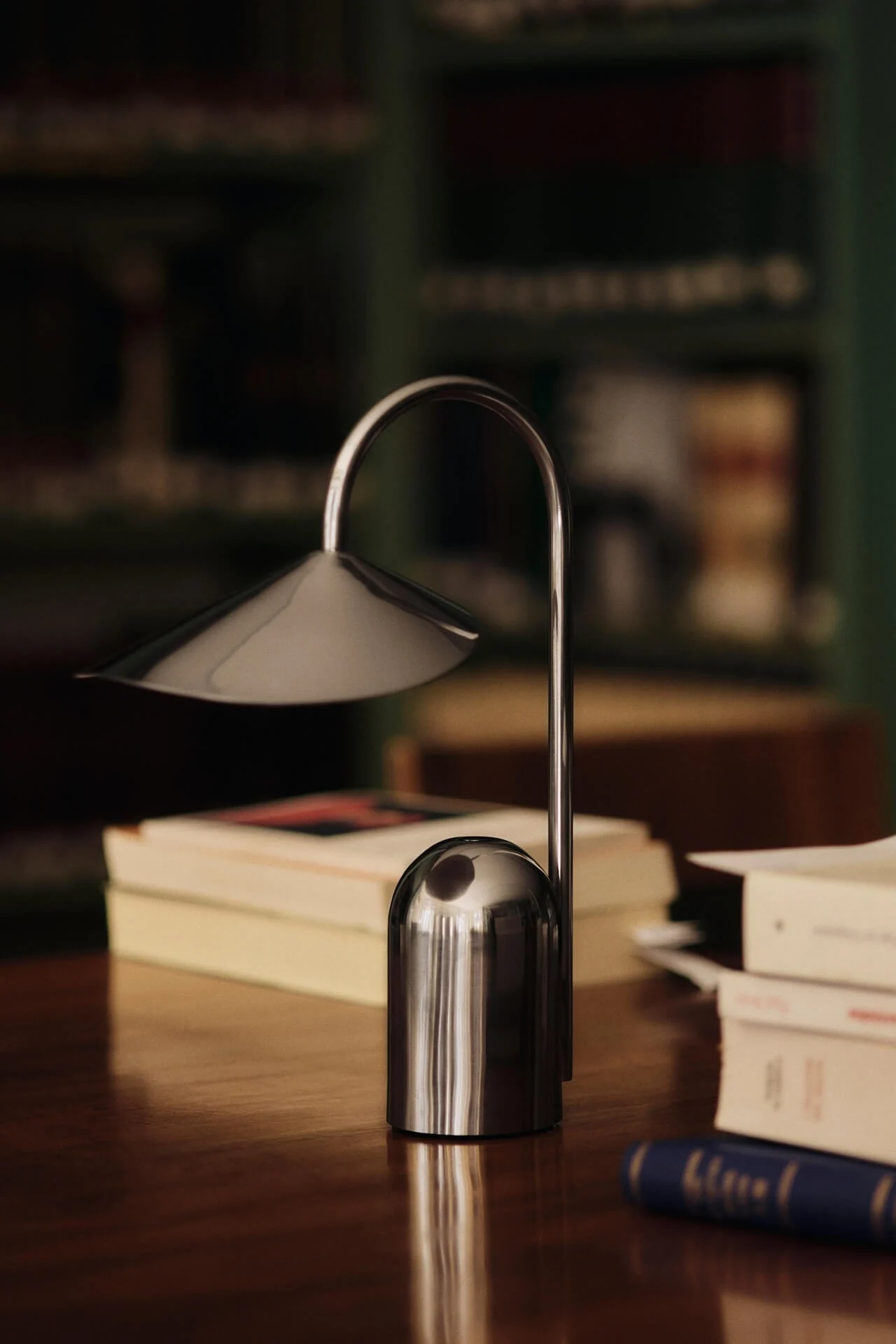 A modern silver table lamp with a curved stem and conical shade sits on a wooden desk with blurred books in the background.