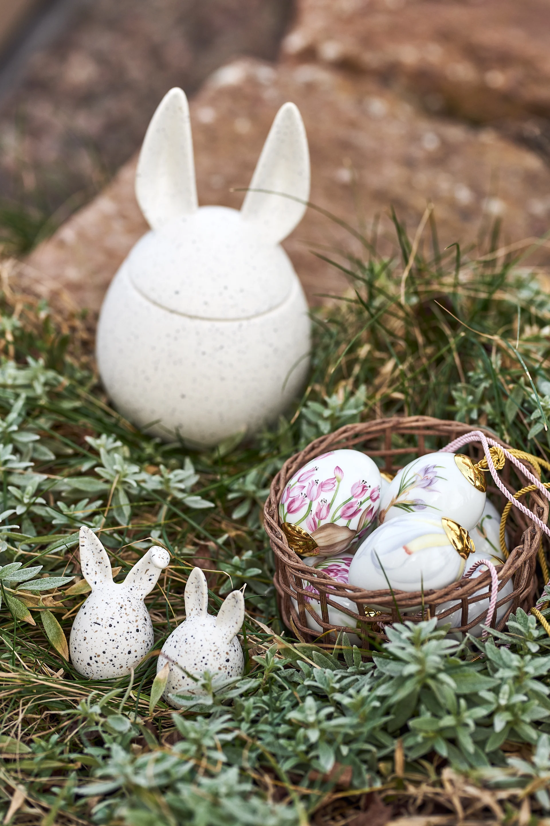 Ceramic bunny container, two small bunnies, and a basket of floral Easter eggs nestled in green grass.