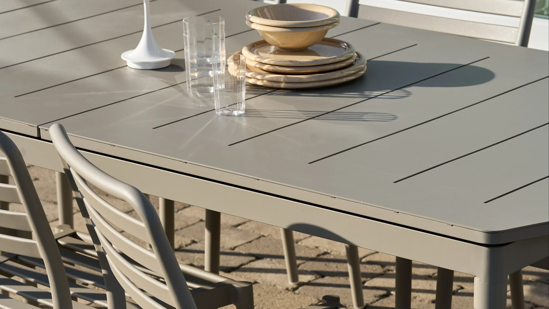 Light grey slatted outdoor dining table with stacked beige plates and bowls, two clear glasses, and a white decorative item.