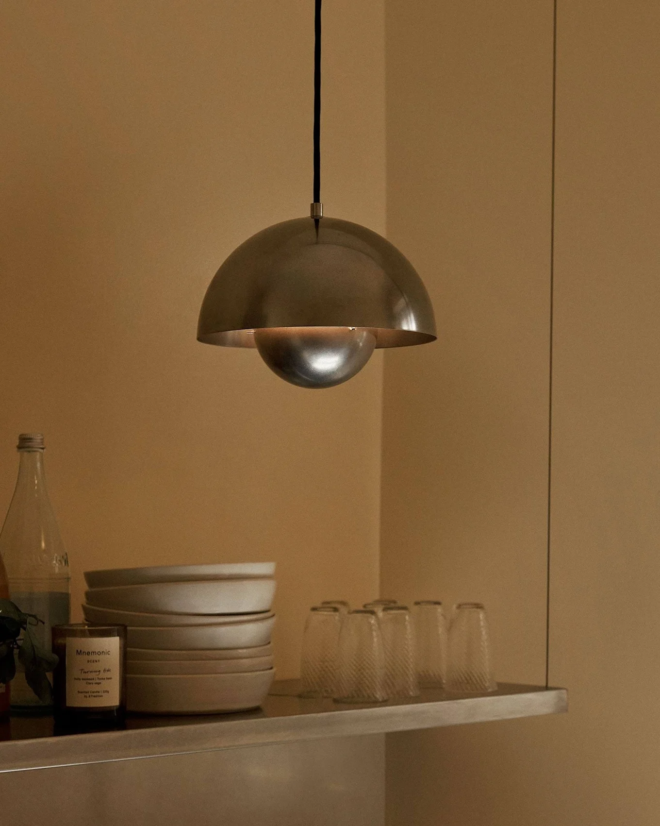 A metal dome-shaped pendant lamp hangs over a shelf with bowls, glasses, bottles, a fruit in a bowl, and a scented candle.