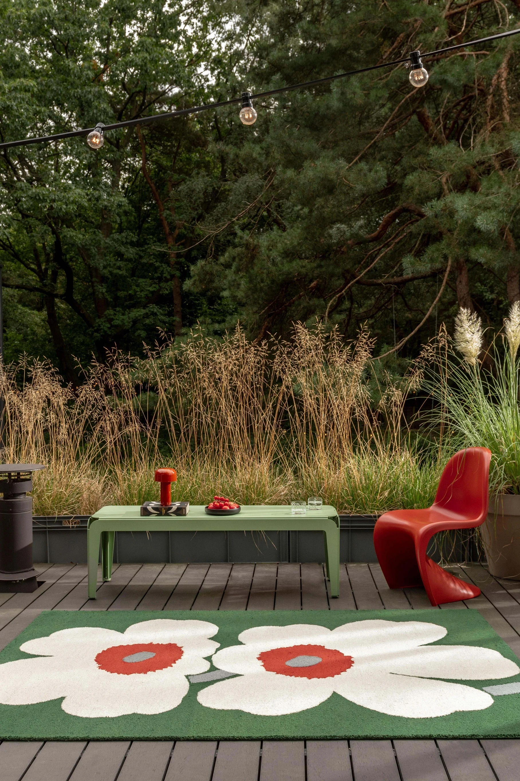An outdoor patio with a green floral rug, a light green table, a red chair, and string lights, surrounded by tall grasses and trees.