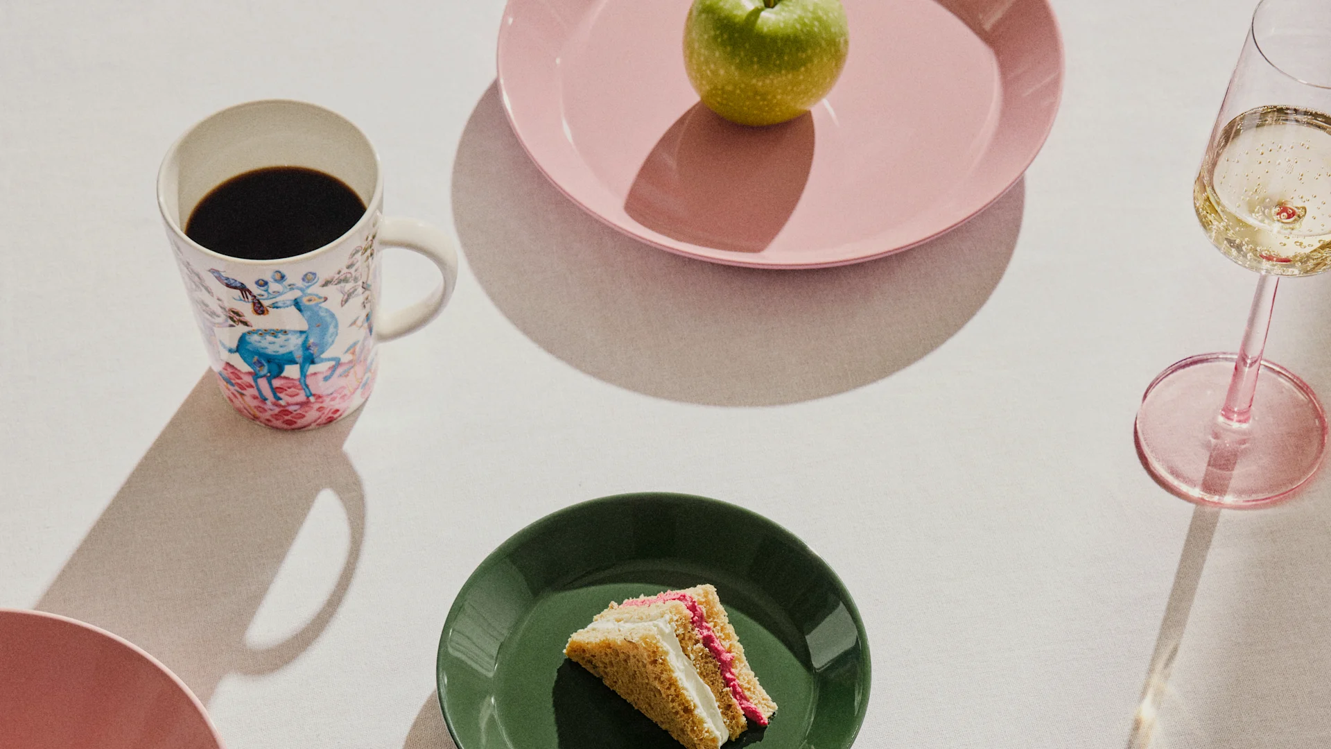 Table setting with a patterned coffee mug, a green apple on a pink plate, a slice of cake on a green plate, and a glass of champagne.