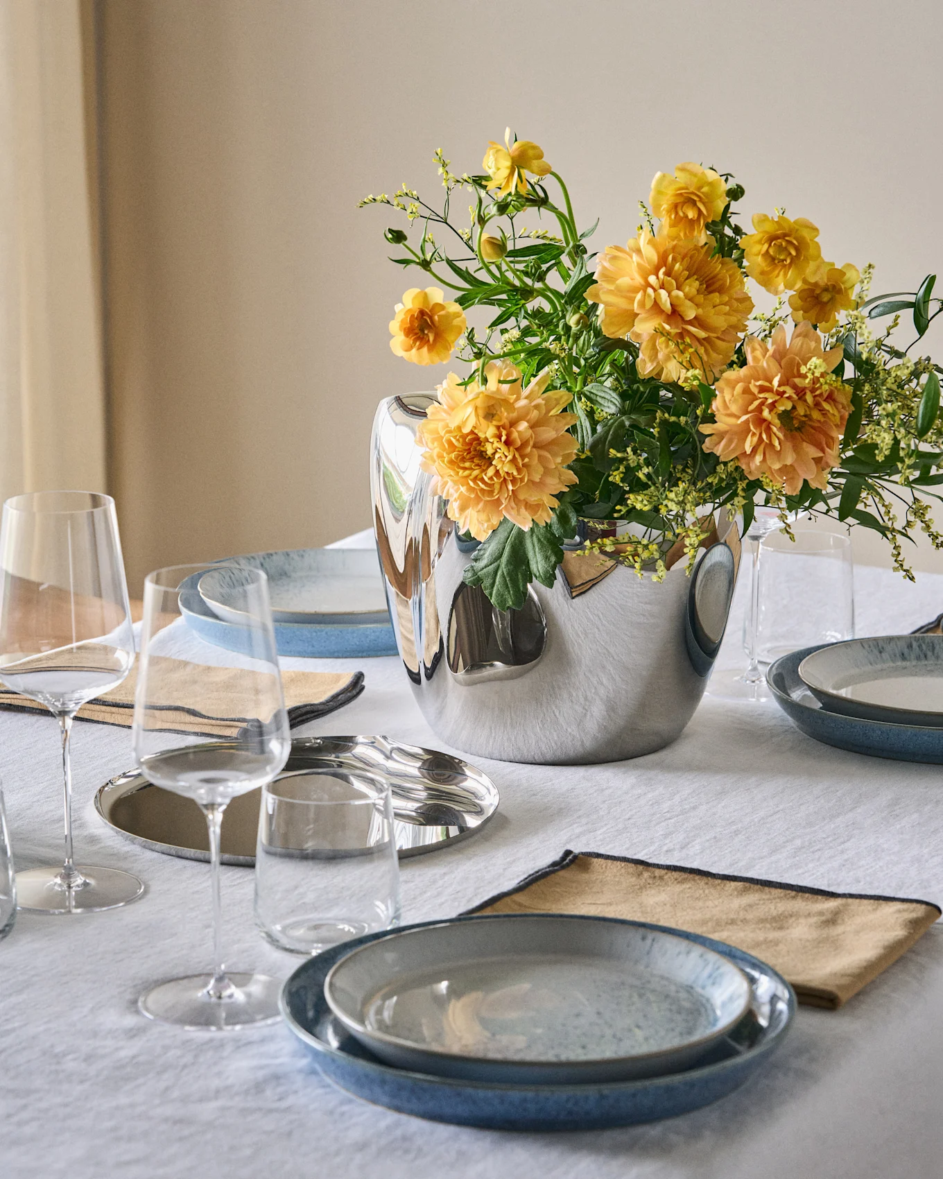 A dining table set with blue plates, tan napkins, clear glasses, and a silver vase holding orange-yellow flowers on a white tablecloth.