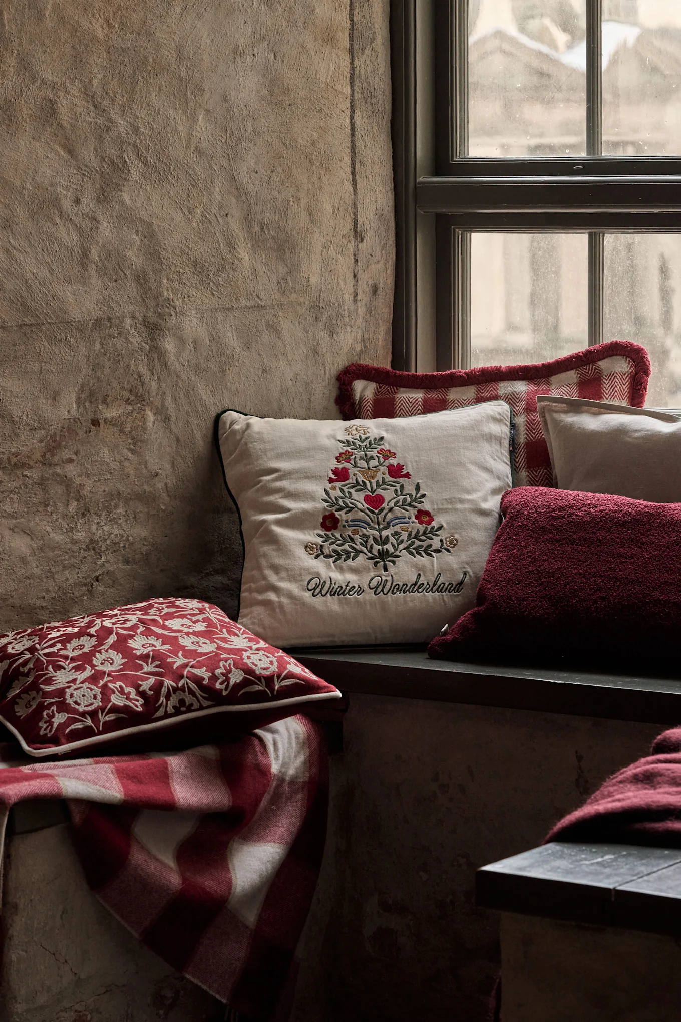 Cozy window nook with festive pillows, including one with an embroidered Christmas tree and 'Winter Wonderland', and a red plaid blanket.