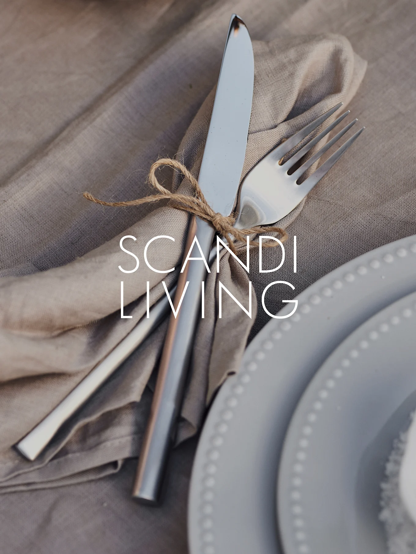 Close-up of knife & fork tied with twine on a linen napkin, next to grey dotted plates. Text: SCANDI LIVING.