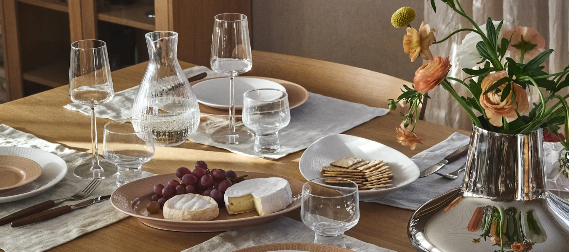 An elegant dining table set with clear glasses, a water carafe, light pink and white plates, cutlery, a cheese and grape platter, crackers, and a silver vase with peach flowers.