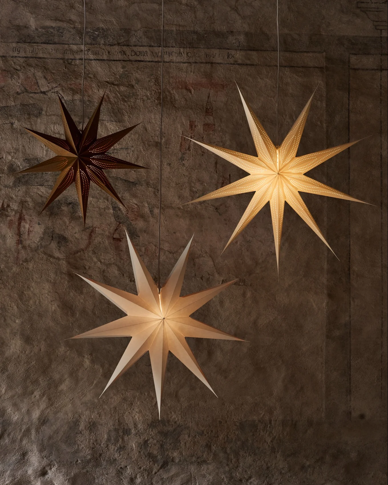 Three star-shaped paper lanterns hang against a textured wall. One dark lantern has tiny perforations, while two brighter ones illuminate the scene.