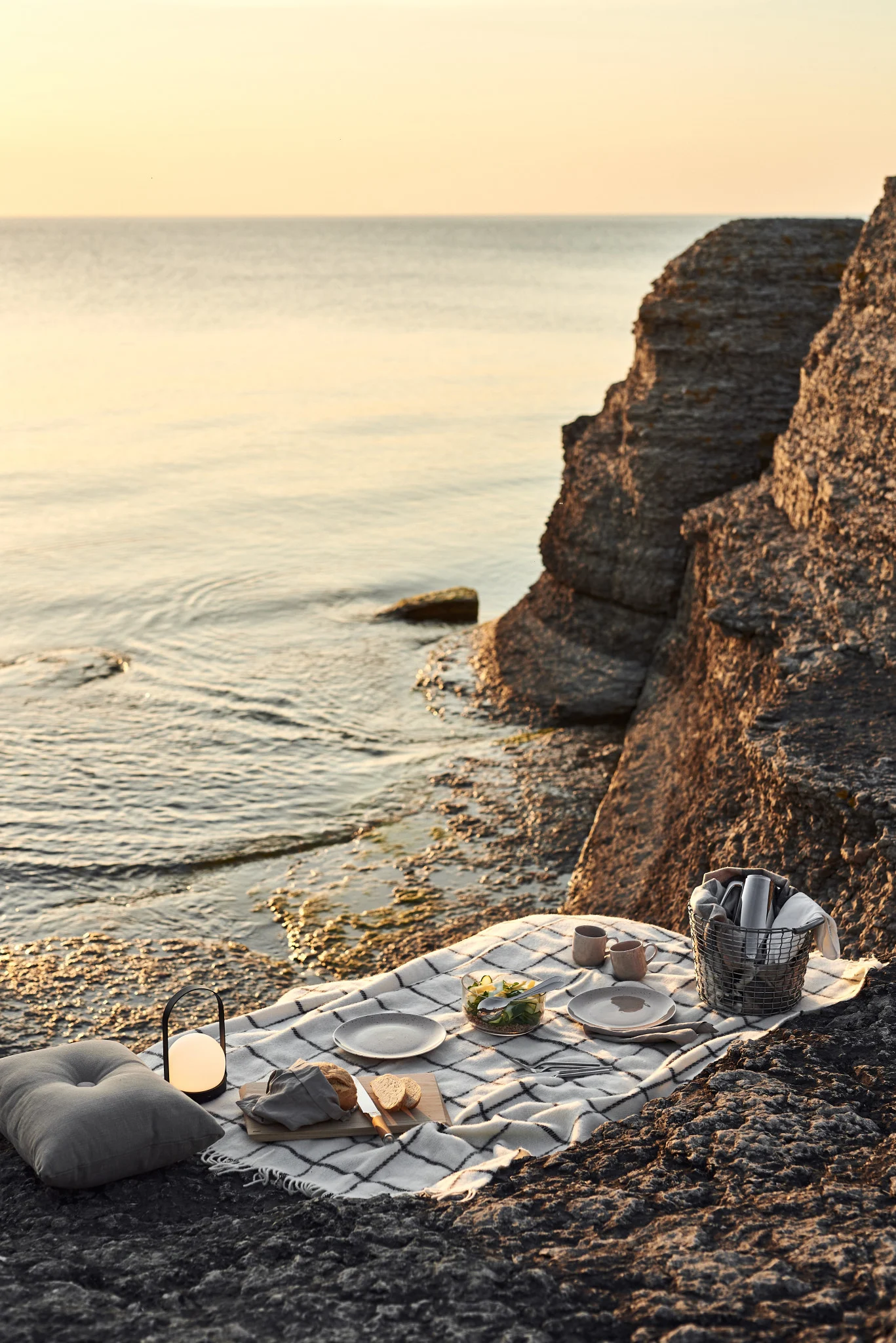 A picnic blanket is laid out on the beach next to the water. The blanket is set with plates, some food a portable lamp and a metal basket to carry the picnic essentials. 