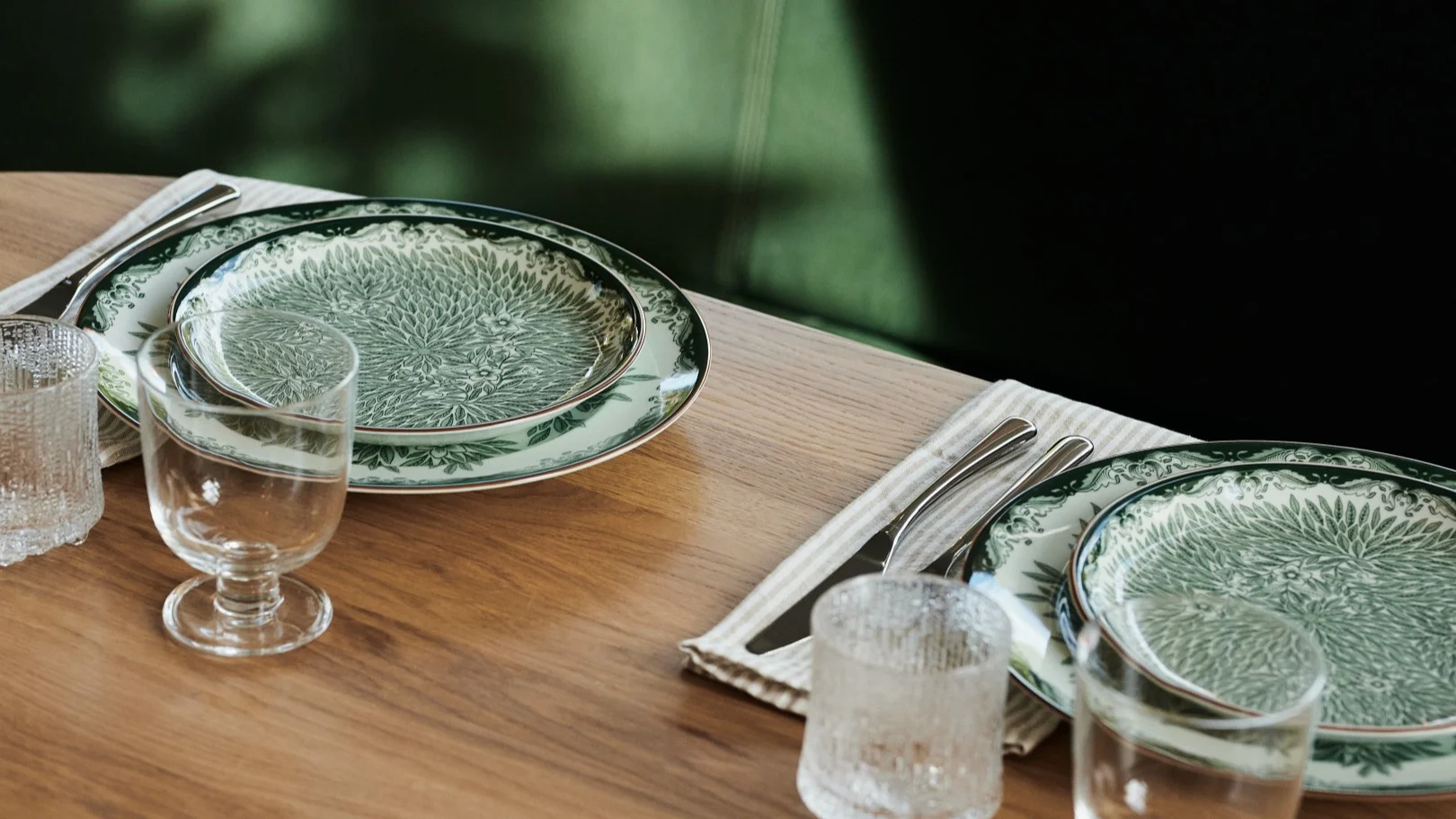 Two place settings on a wooden table, with green patterned plates, glasses, and silverware on striped napkins.
