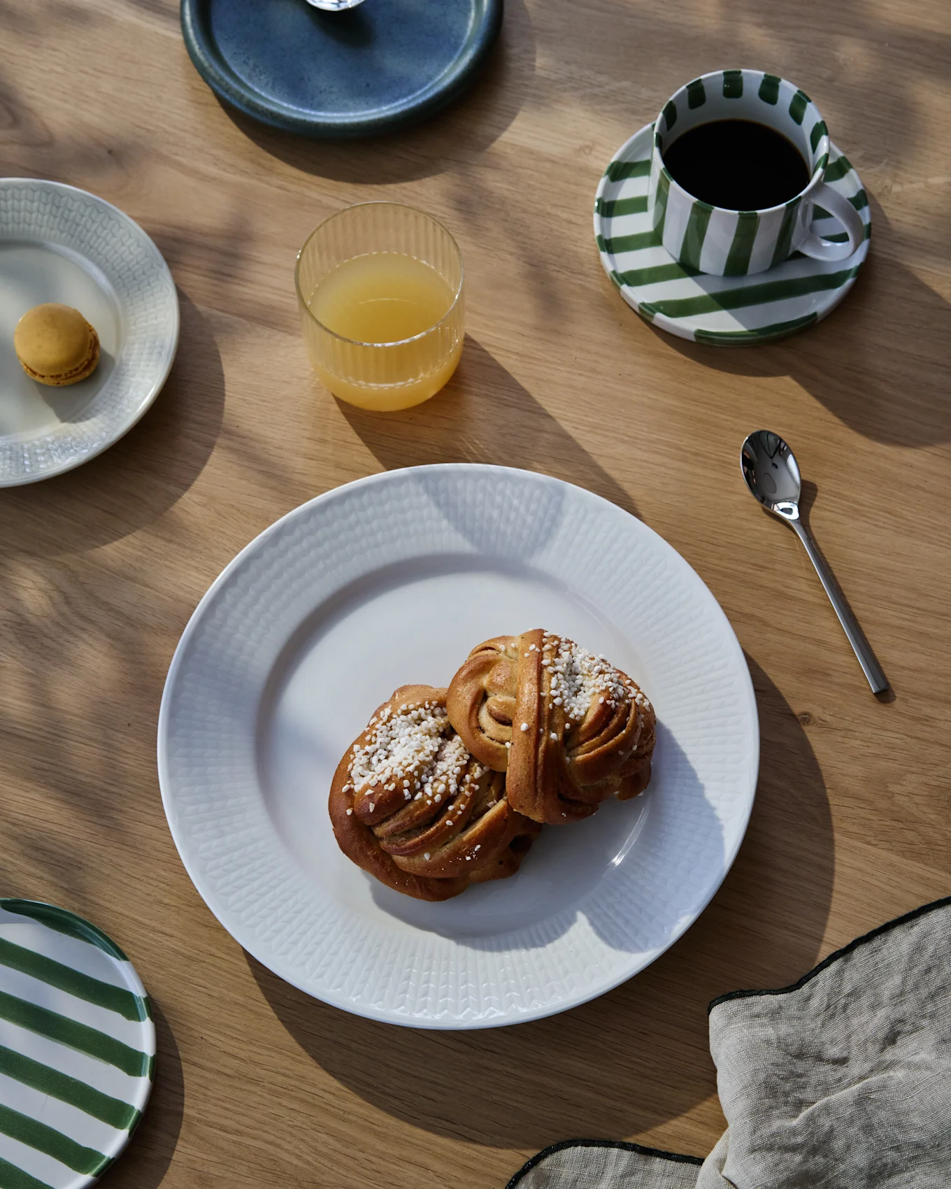 A breakfast spread on a wooden table featuring two cinnamon rolls, coffee in a striped cup, orange juice, and a macaron.