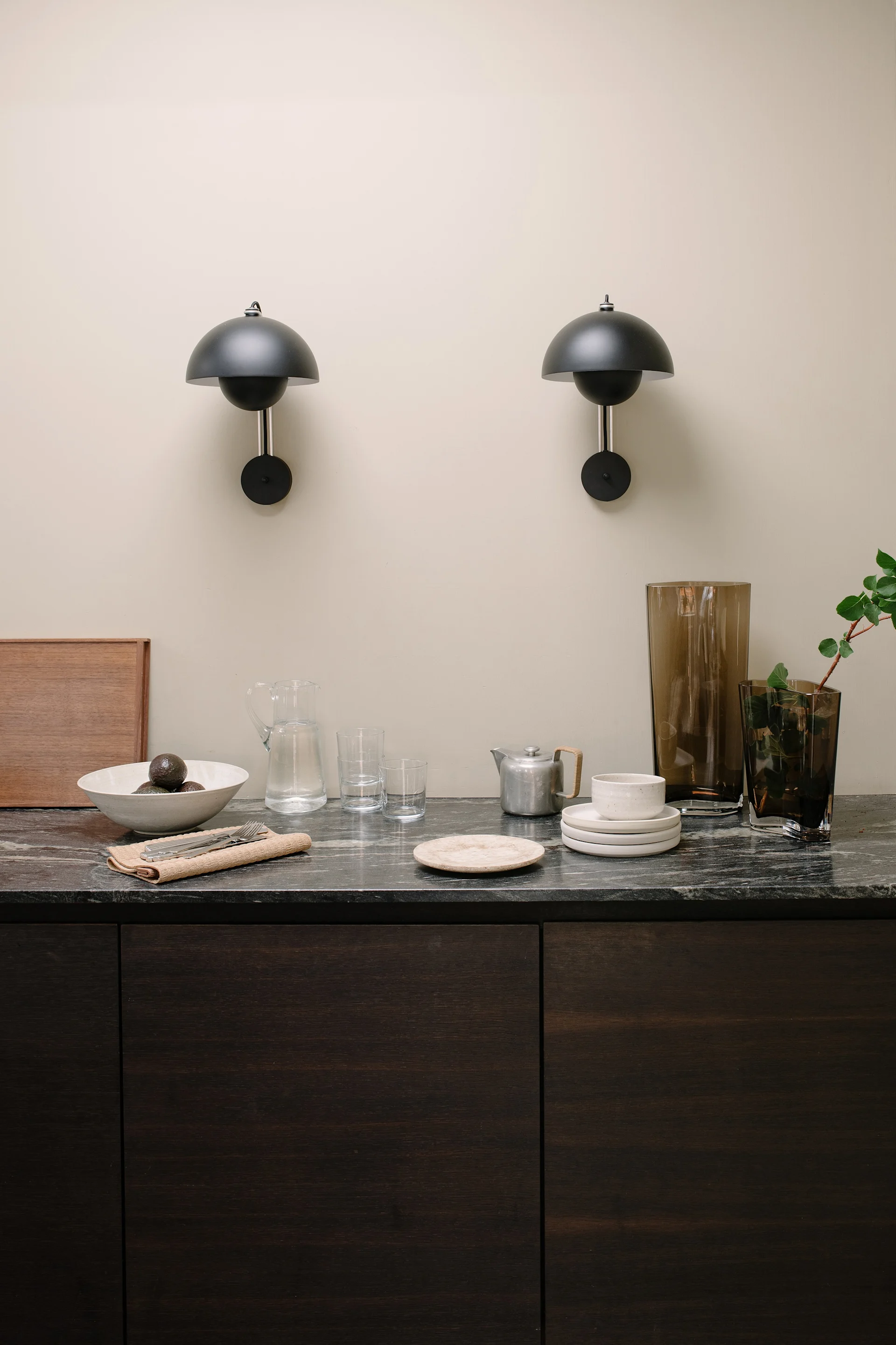 Two black dome-shaped wall lamps above a dark marble countertop with kitchenware, including a bowl of avocados, a teapot, plates, and vases.