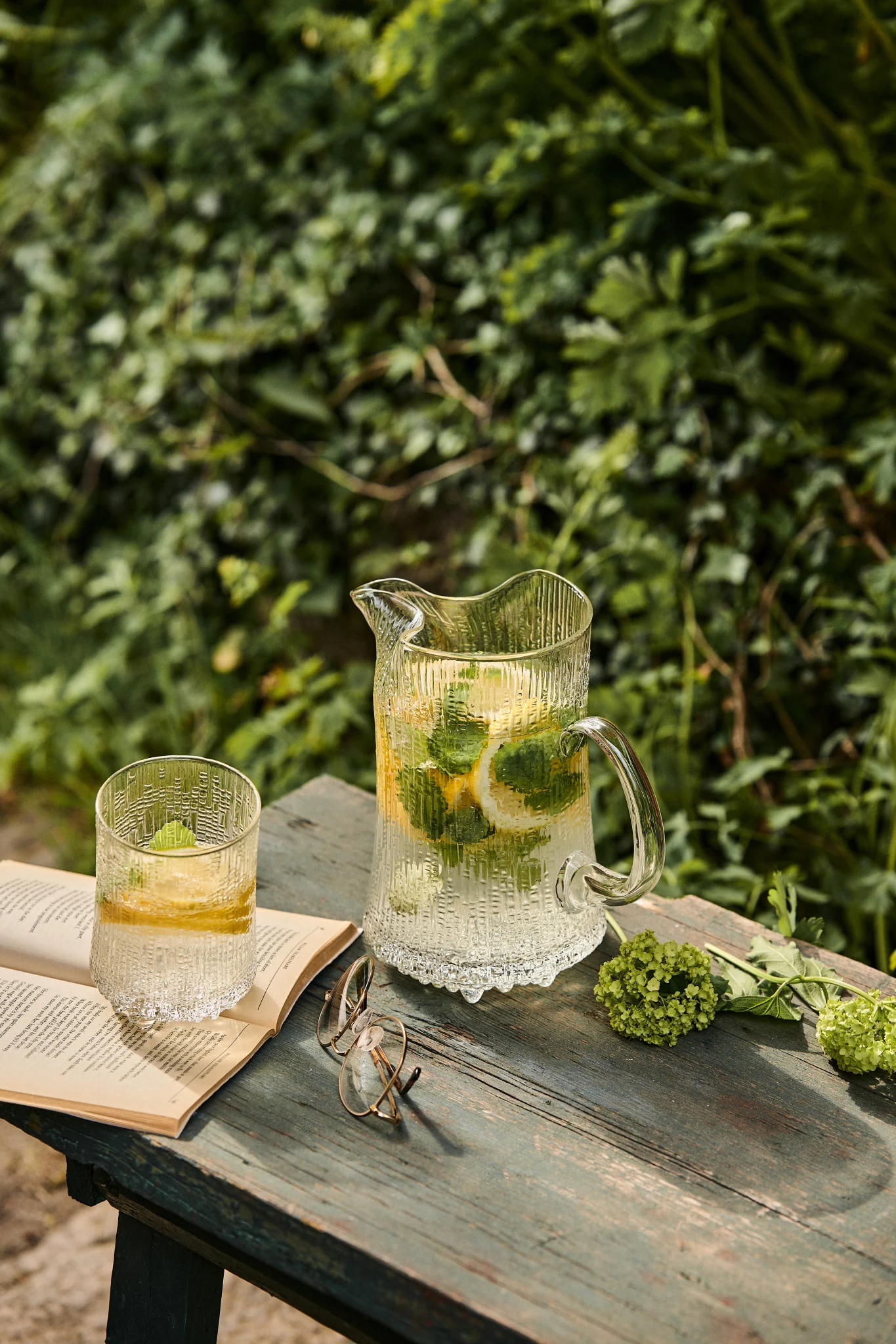 The Ultima Thule jug and matching glass from Iittala stand on a bench in the sunshine with a book. Making your own elderflower cordial is essential for any summer bucket list ideas. 