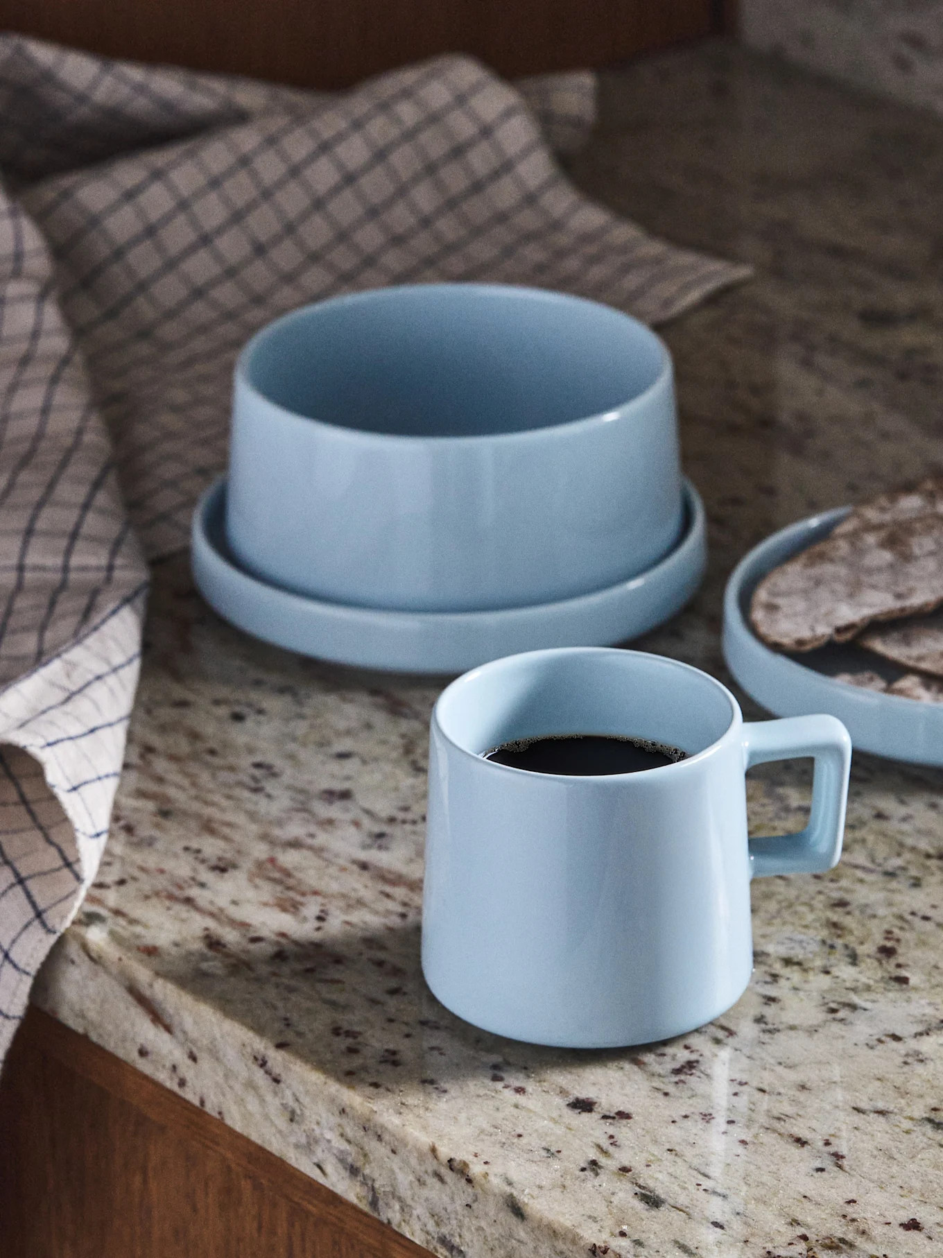 Light blue ceramic coffee mug, bowl, and plate with flatbread on a granite countertop.