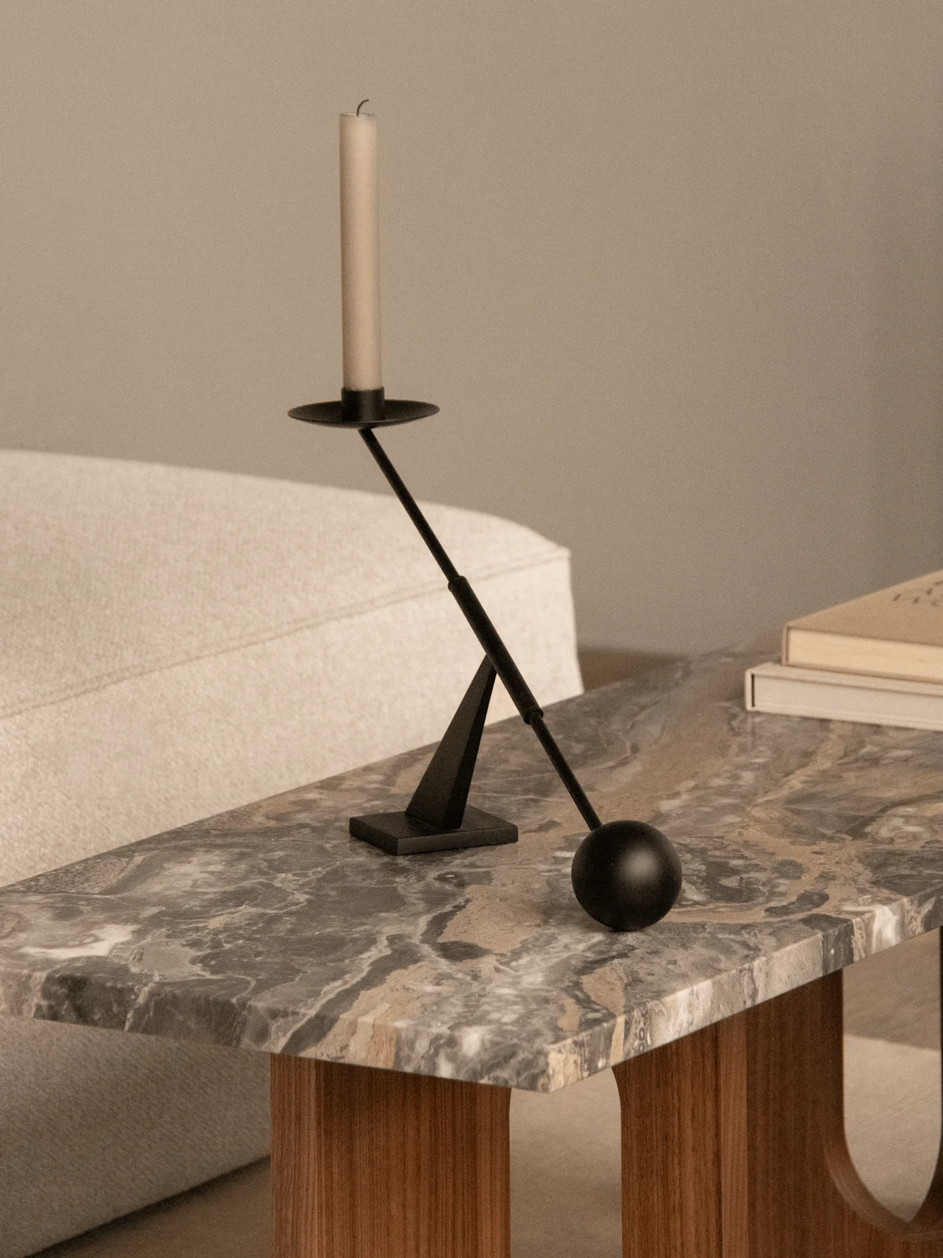A black modern candlestick with a white candle on a marble coffee table next to two books and a light-colored sofa.