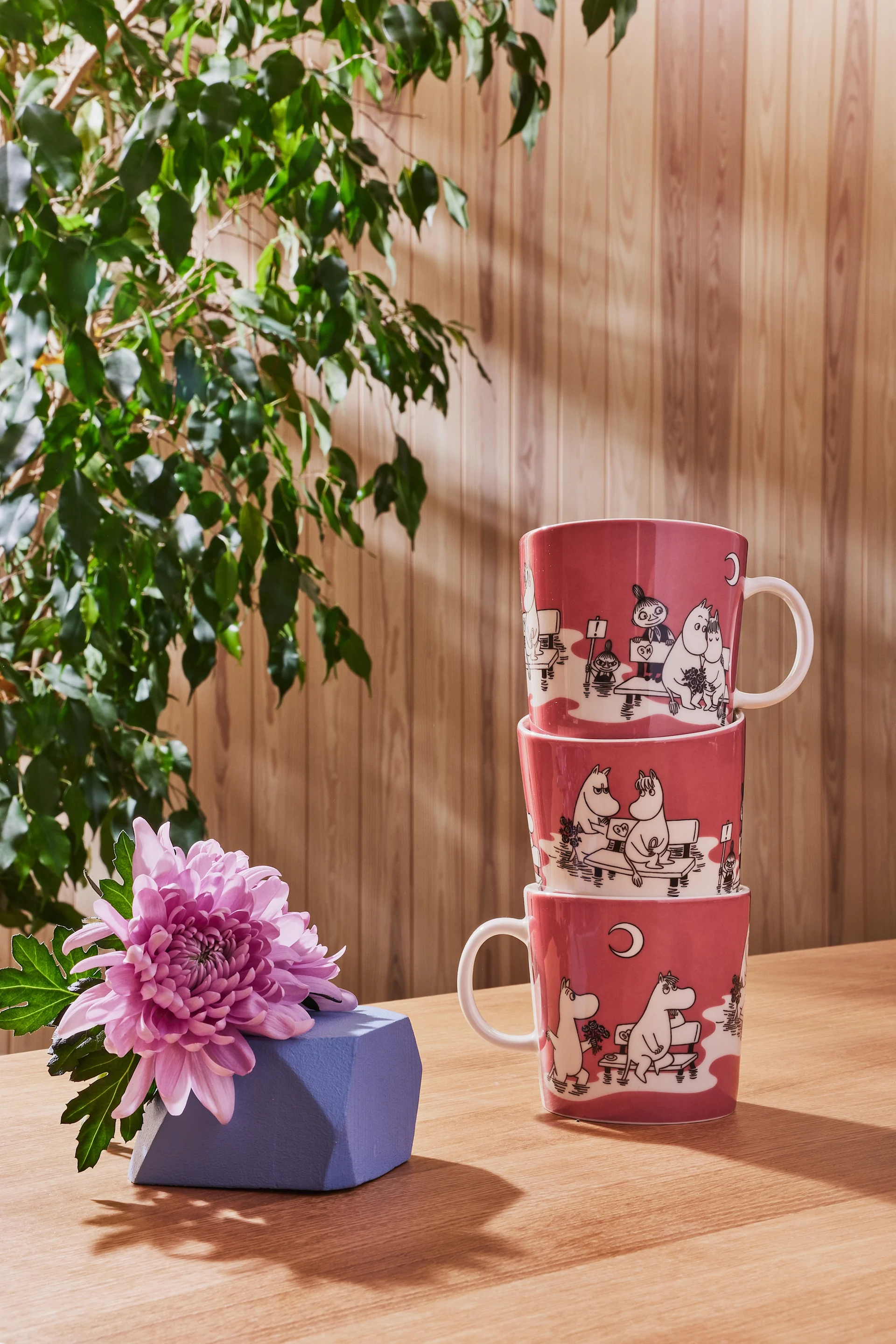 Three pink Moomin mugs from the colour collection are stacked on a table, with a gift lying next to them.