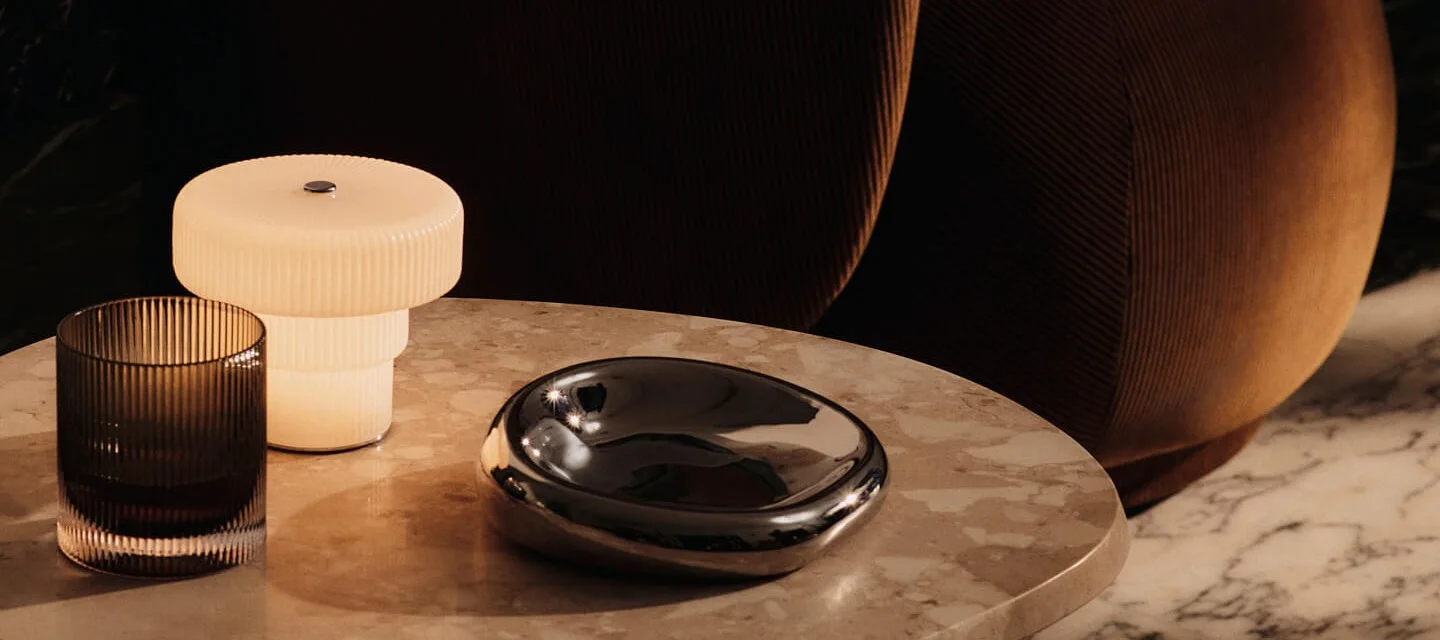 A ribbed table lamp, dark glass, and a shiny decorative bowl on a marble table.