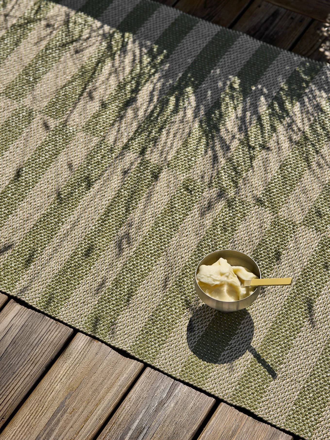 A silver bowl of dessert with a yellow spoon sits on a green and beige striped outdoor mat on a wooden deck, dappled by palm tree shadows.