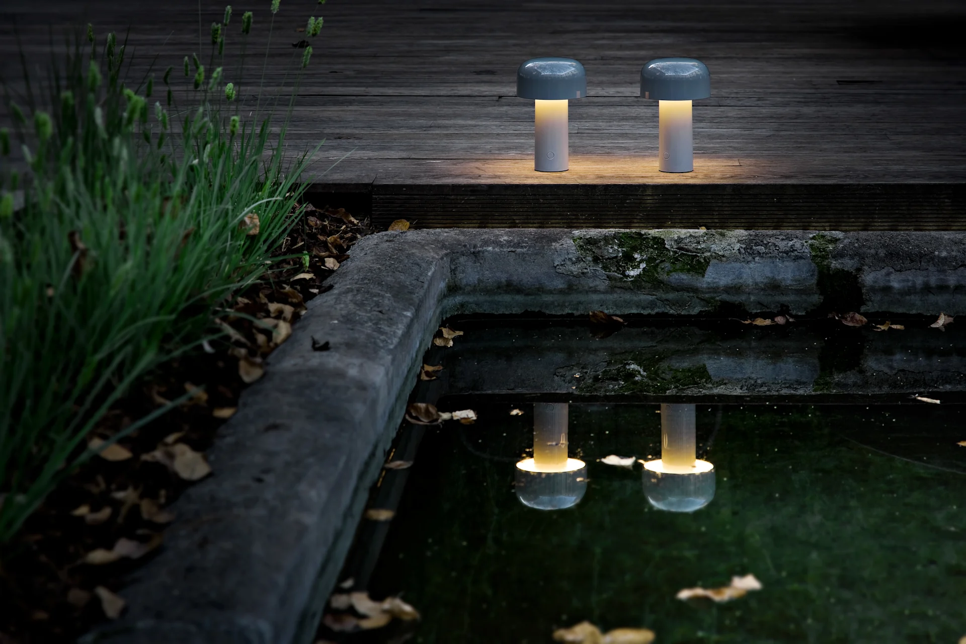 Two portable mushroom-shaped table lamps with warm yellow light sit on a dark wooden deck by a pond, reflecting their glow.