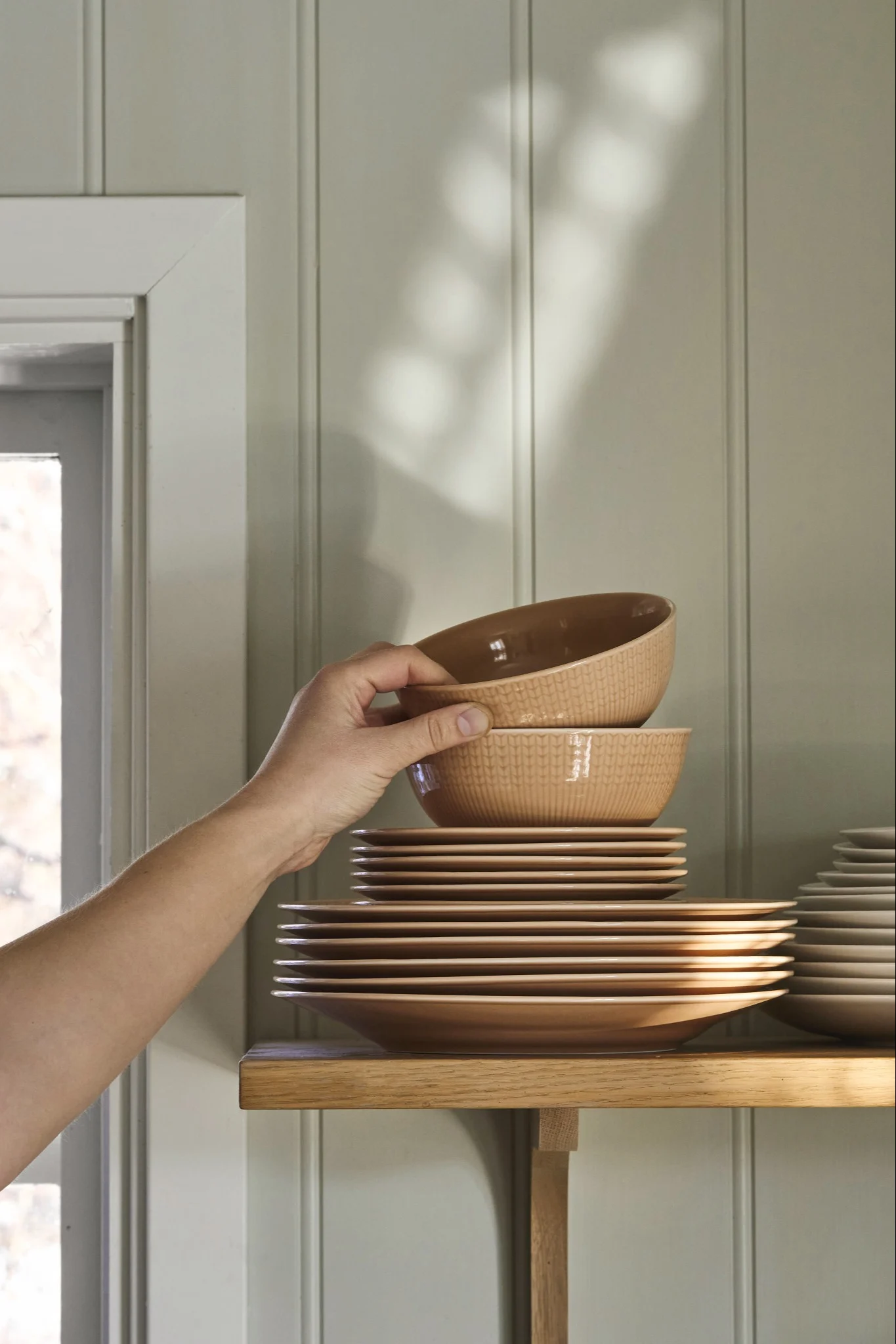 Hand placing a light brown, ridged bowl onto a stack of matching bowls and plates on a wooden shelf.