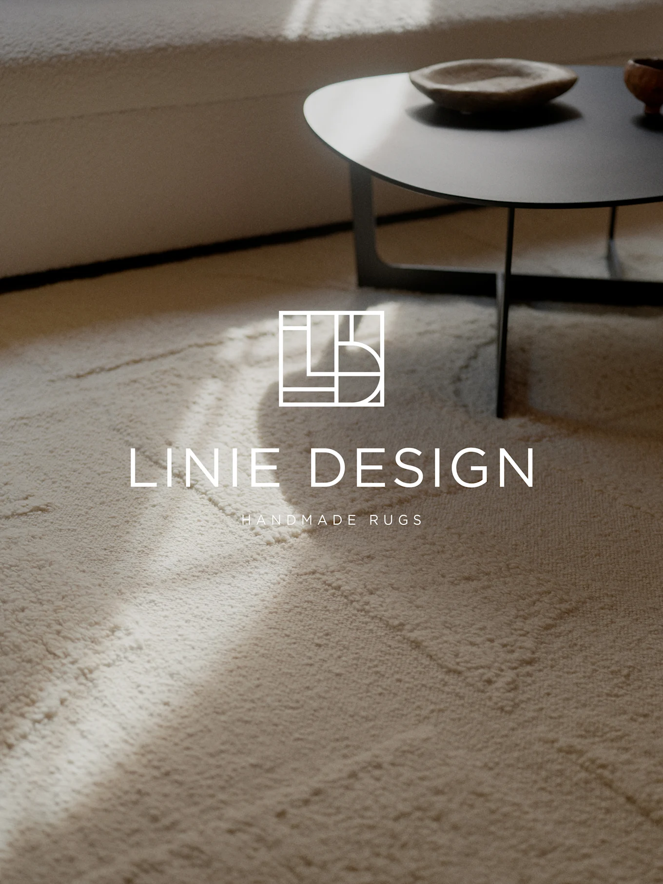 A light textured rug with the Linie Design logo and text 'Handmade Rugs'. A sofa and coffee table are blurred in the background.
