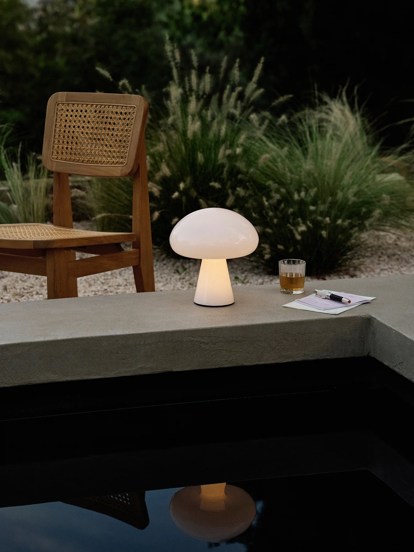 A glowing mushroom-shaped lamp on a concrete ledge next to a pool, with a wooden chair, drink, and pen.