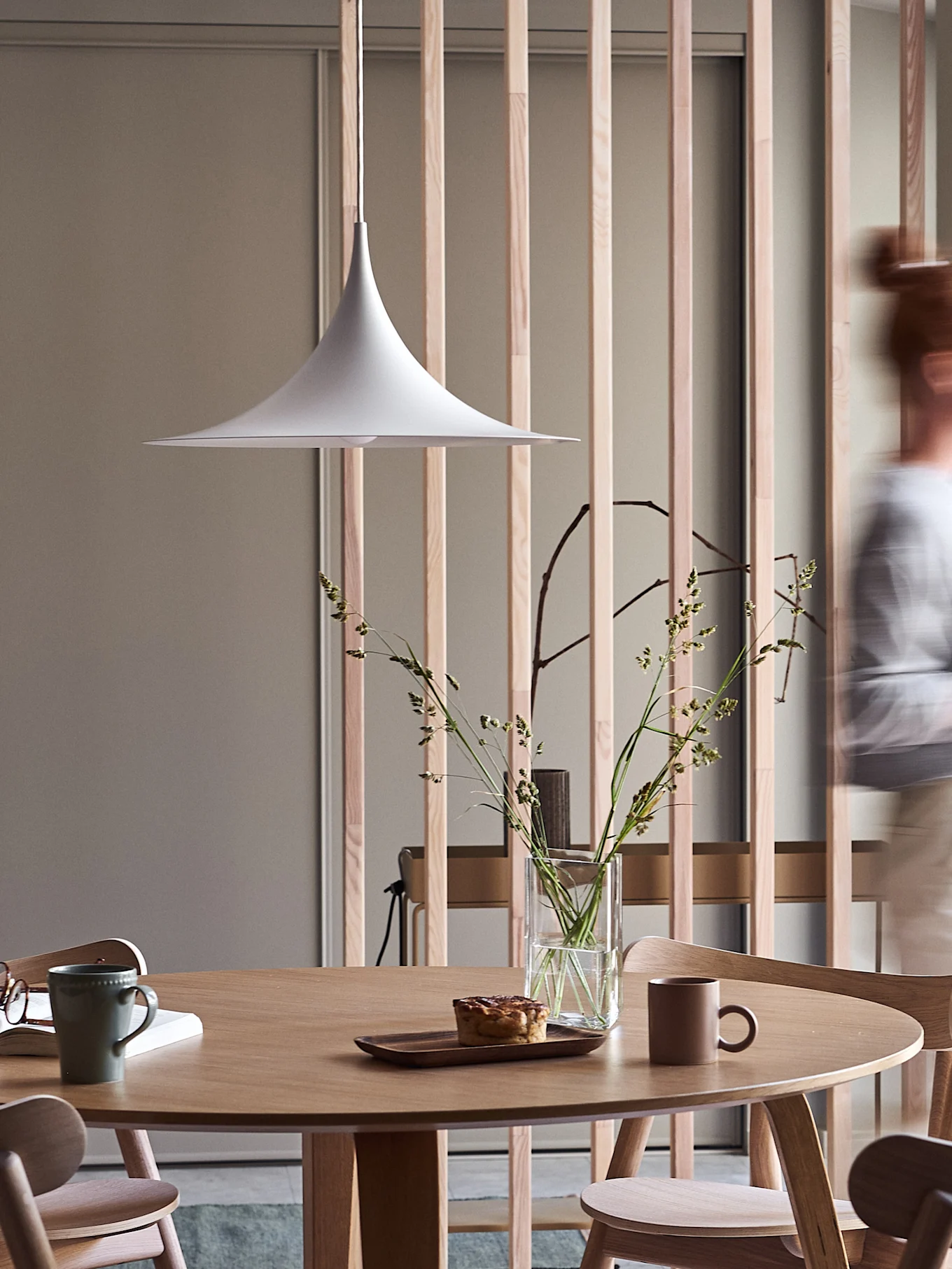 Modern dining area with round wooden table, white pendant lamp, vase of grasses, pastry, and mugs, with a blurred person walking by.