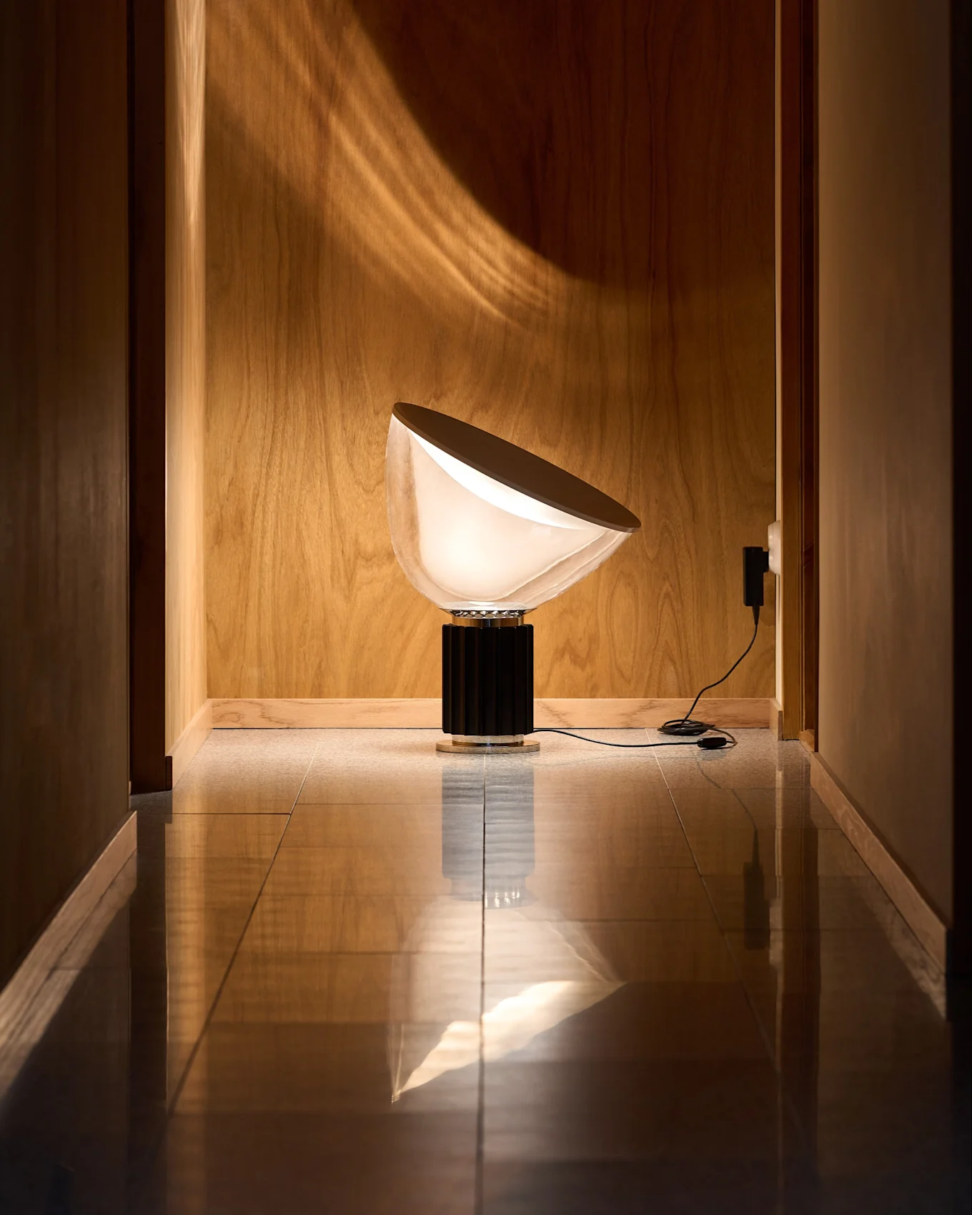 Modern lamp with a semi-circular, white opaque shade and black ridged base in a wood-paneled hallway with a reflective floor. Light pattern on wall.