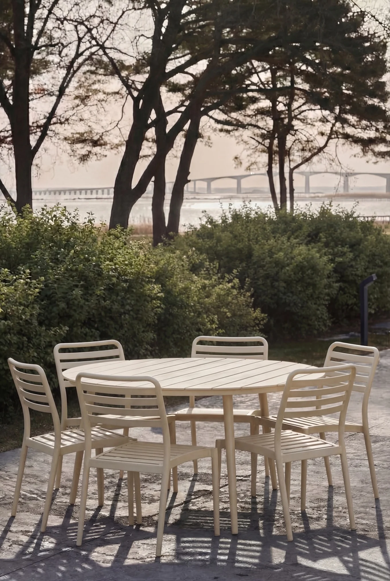Light-colored outdoor dining set with a round slatted table and six matching chairs on a patio with trees and a bridge in the background.
