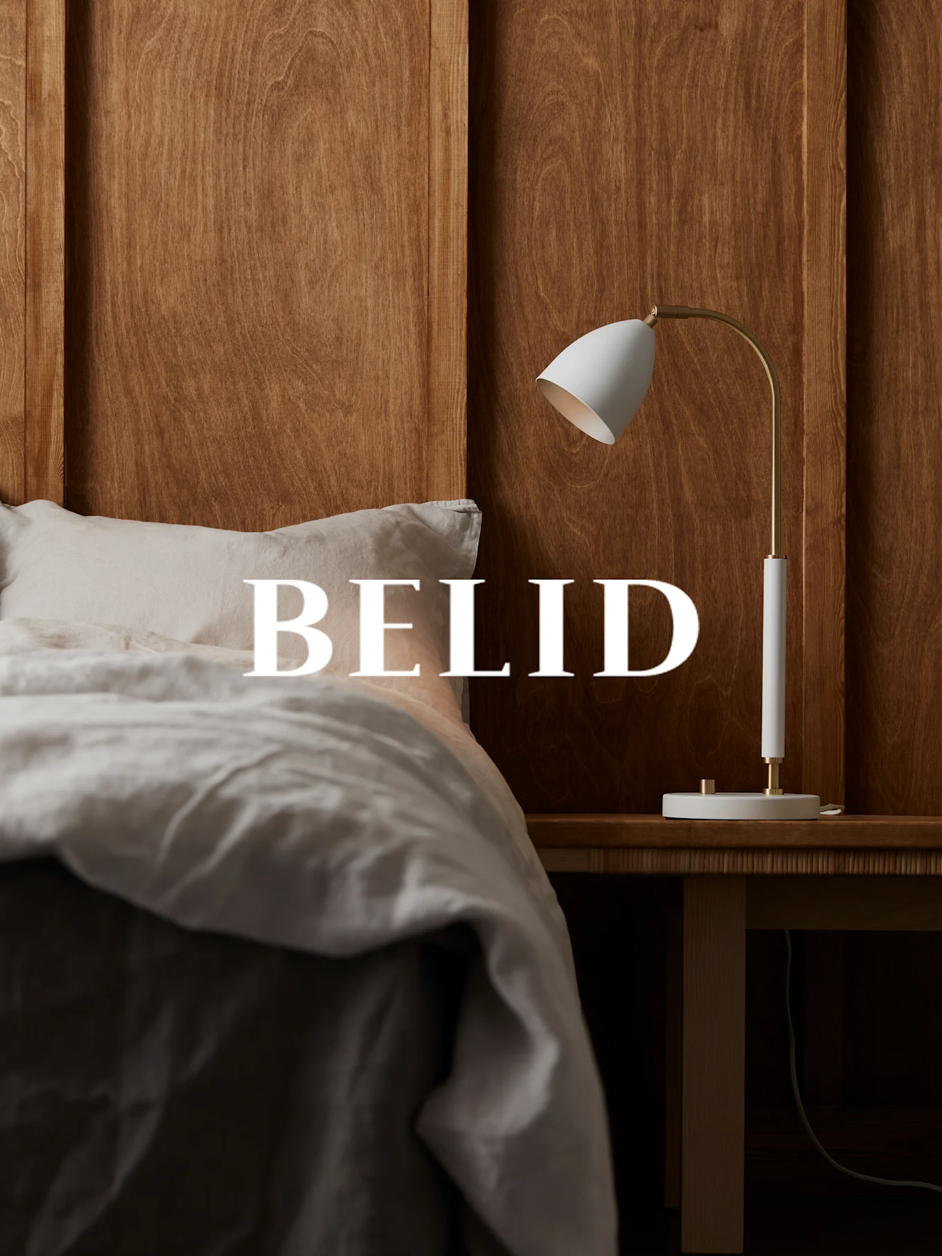 White and brass lamp on a wooden bedside table, with 'BELID' text over a bed with linen sheets.