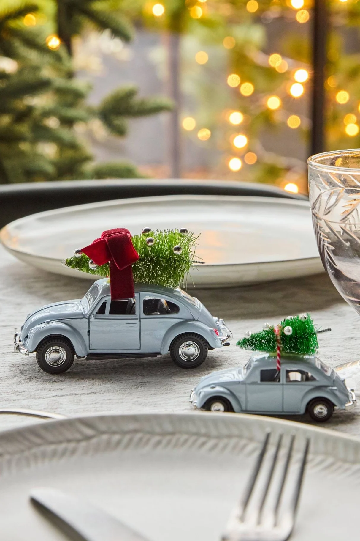 Two light blue toy cars with miniature Christmas trees, one with a red bow, sit on a festive dining table with blurred holiday lights.