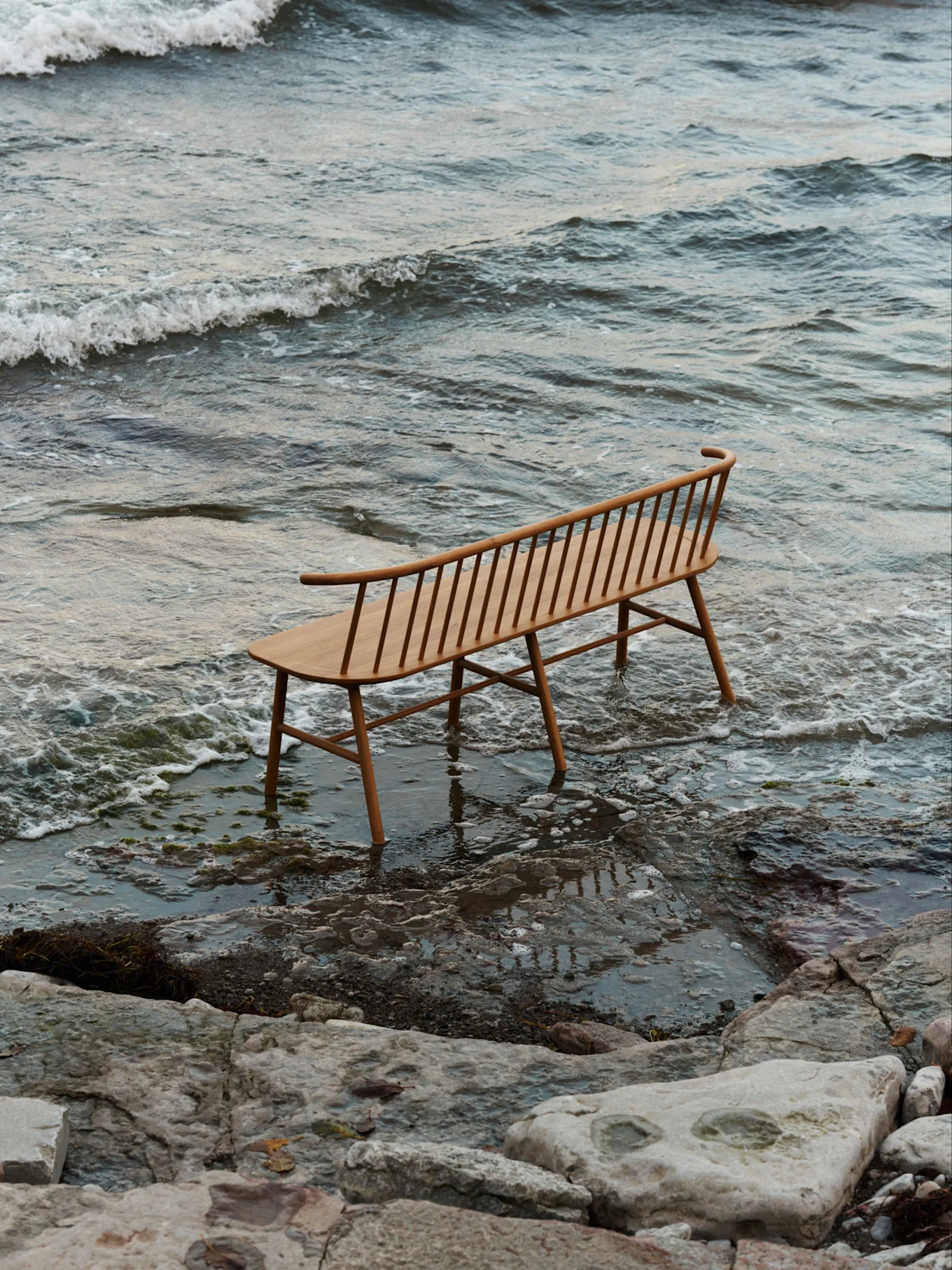 A light wooden slatted bench with a curved backrest stands in shallow water on a rocky beach with gentle waves.