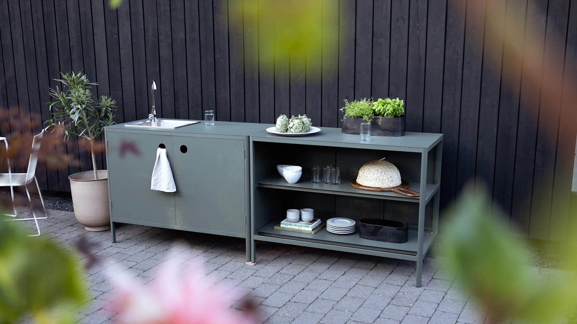 Olive green outdoor kitchen unit with sink, open shelves, potted herbs, dishes, and a woven food cover on a patio.