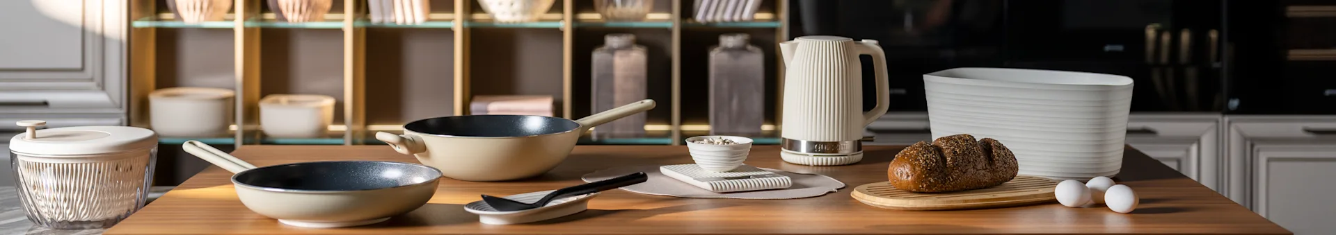 Kitchen counter with beige frying pans, a white ribbed kettle, a seeded bread loaf on a board, and eggs. Modern kitchen accessories.