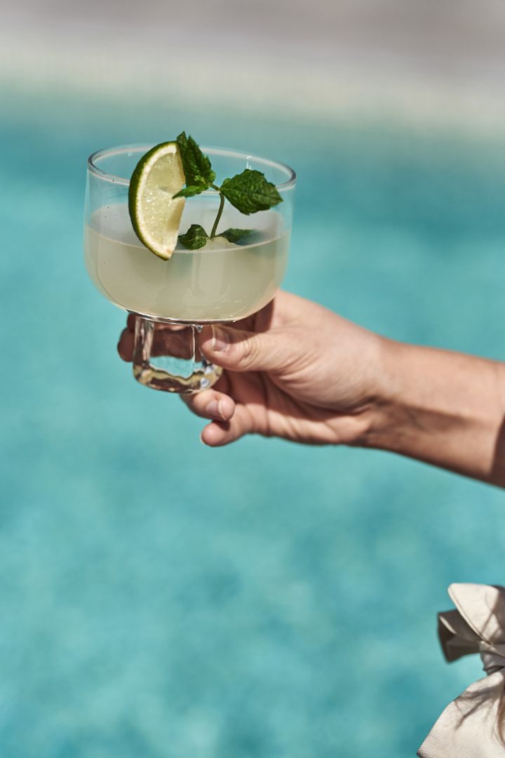 Discover these refreshingly simple summer cocktails and mocktails. Here you see a hand hold a mojito with a wedge of lime next to a pool. 