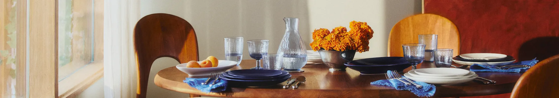Round wooden dining table with blue and white plates, blue napkins, and orange flowers. Sunlight filters from a window.