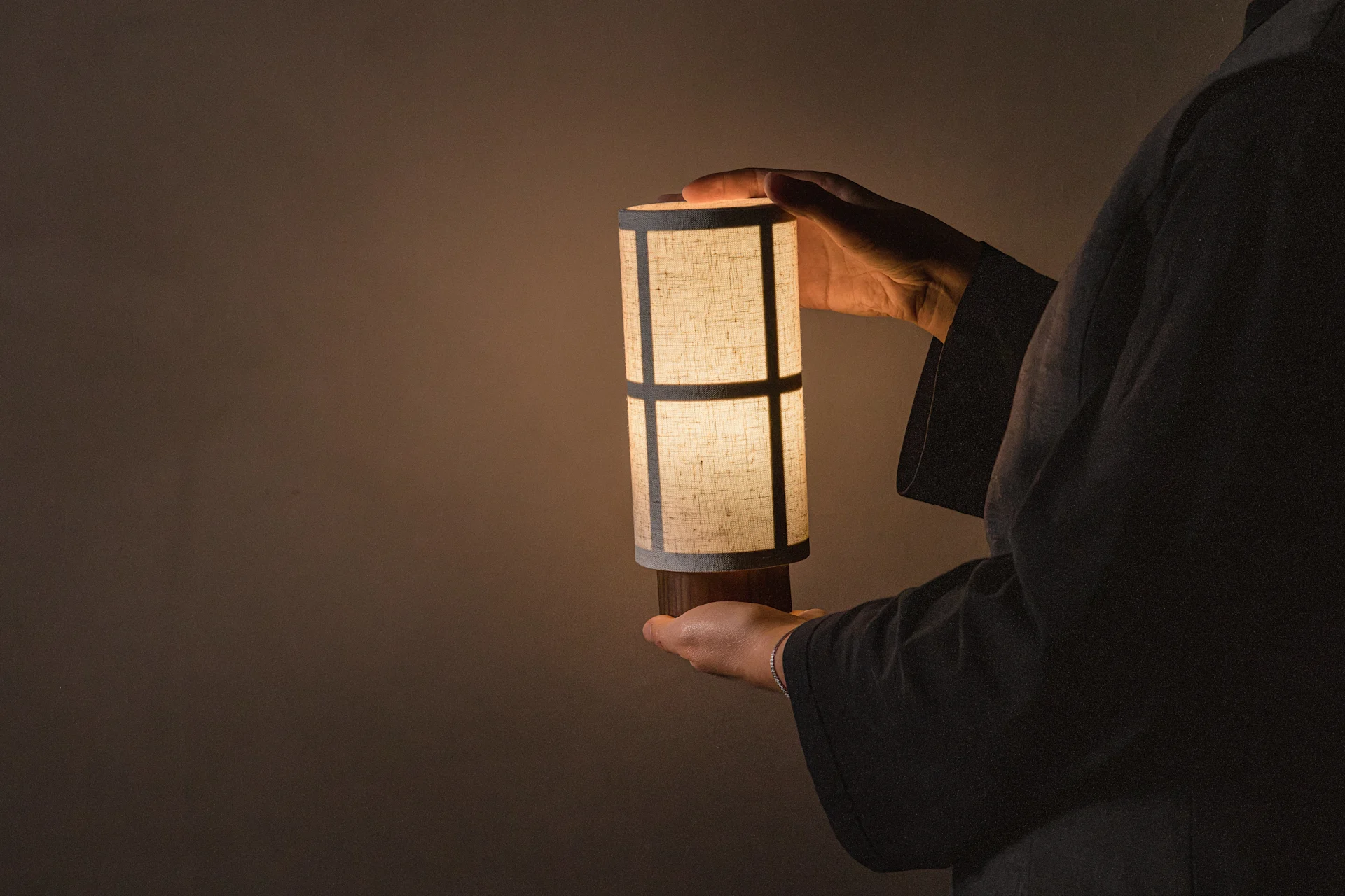 Person holding an illuminated cylindrical table lamp with a patterned fabric shade, casting a warm glow.