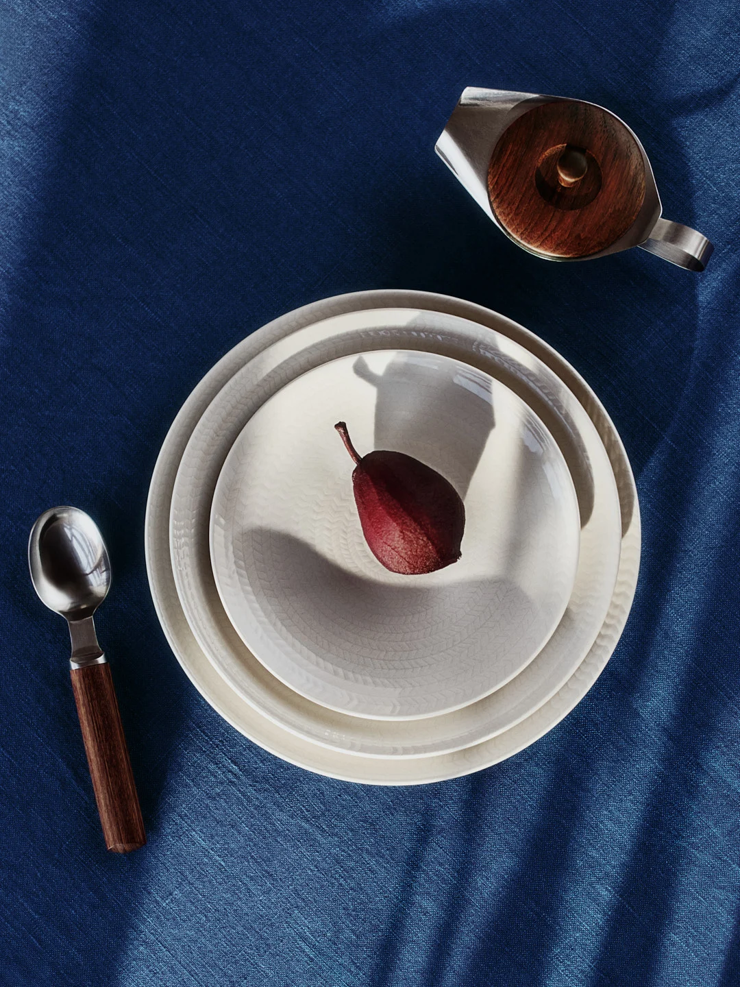 Red pear on two patterned plates, spoon, and creamer on blue tablecloth with shadows.