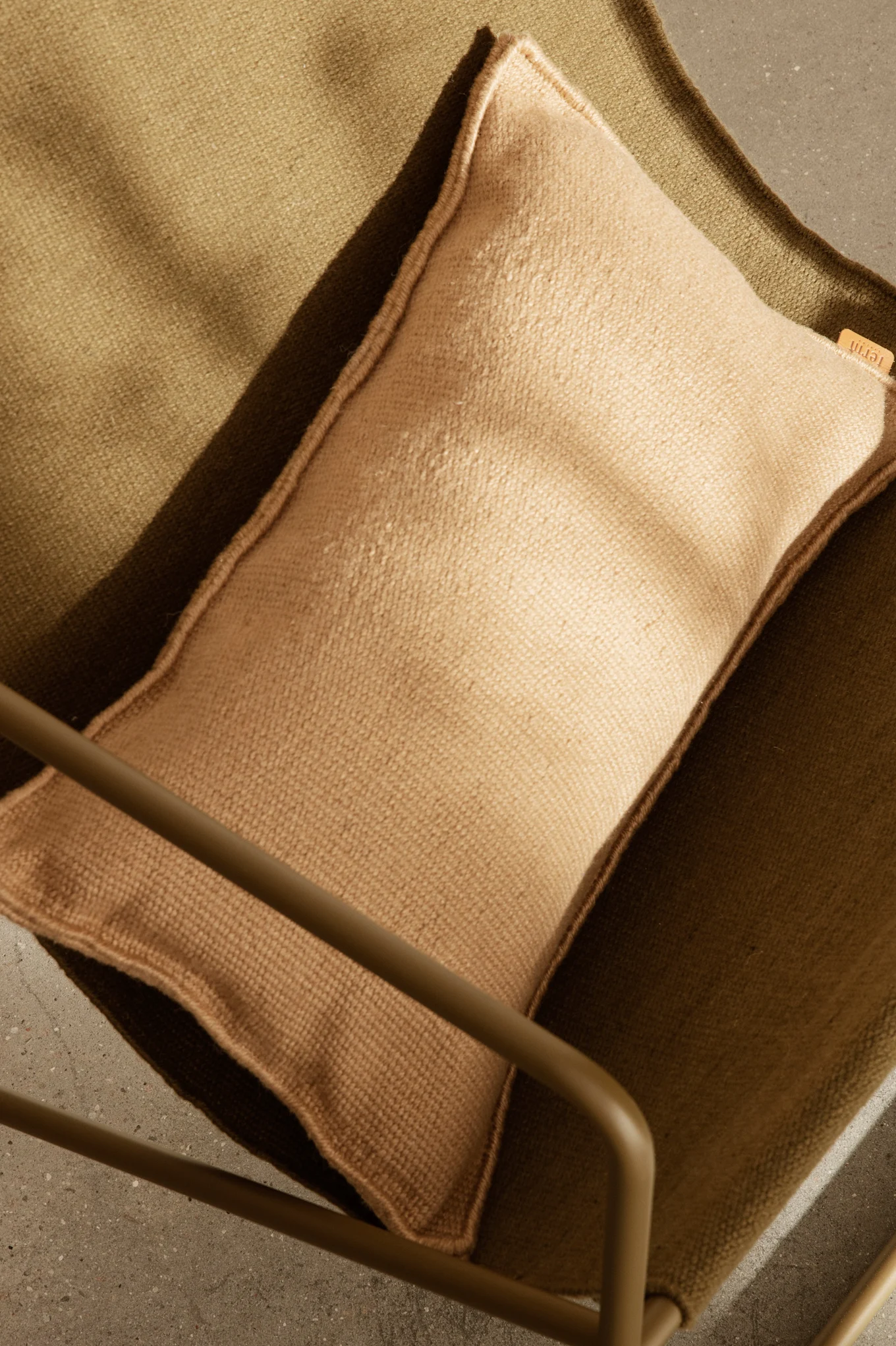 Overhead view of a light brown cushion on a dark olive green textured fabric chair, with part of a metal frame and concrete floor.