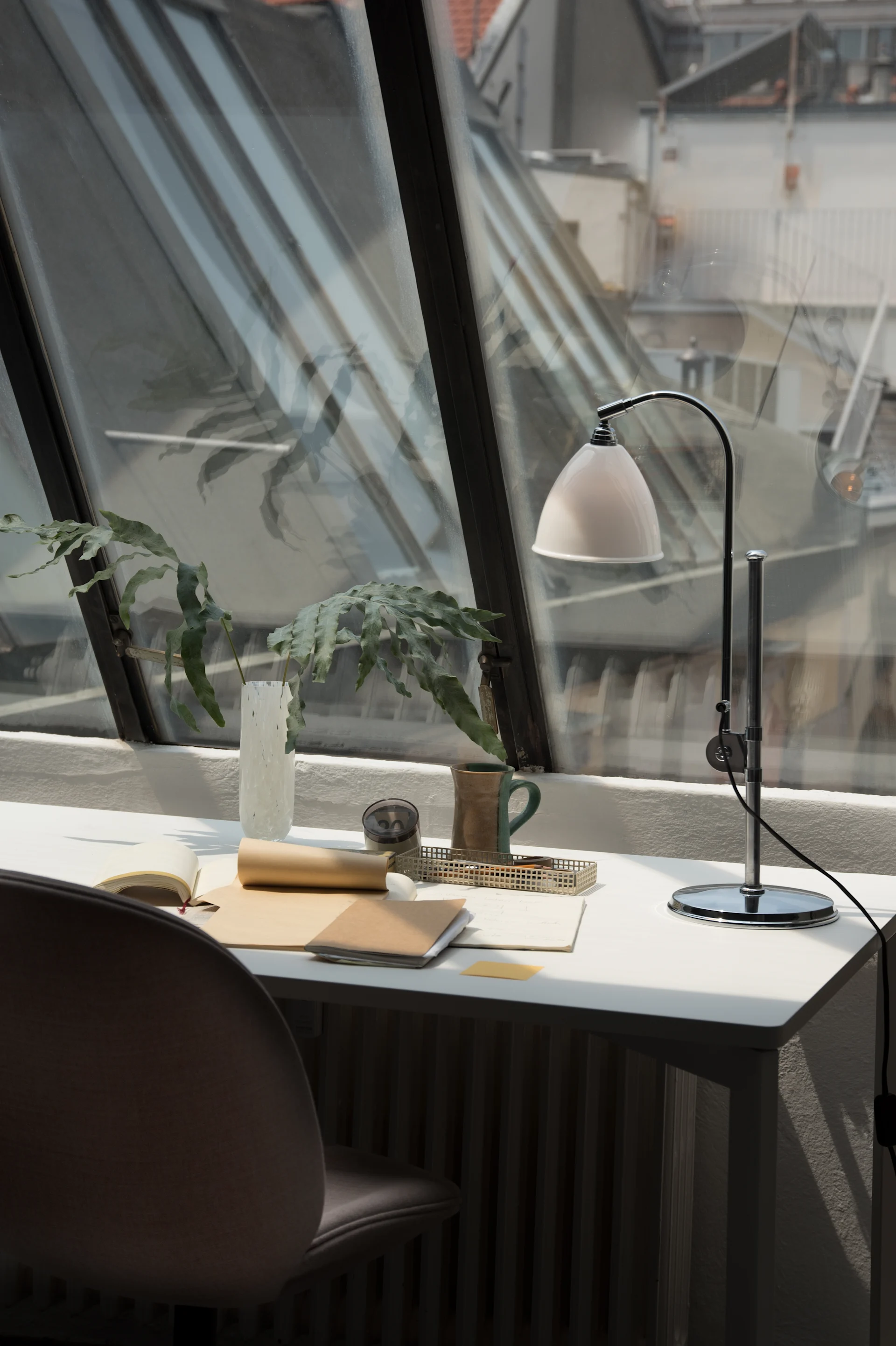 A modern desk setup by a slanted window overlooks rooftops, featuring a white lamp, green plant, notebooks, and a two-toned mug.