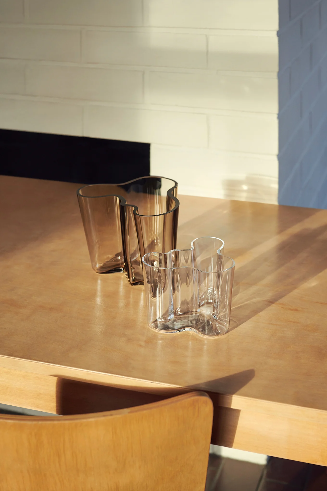 Three wavy glass vases on a wooden table. Two are amber, one is clear, with sunlight and shadows across the scene.
