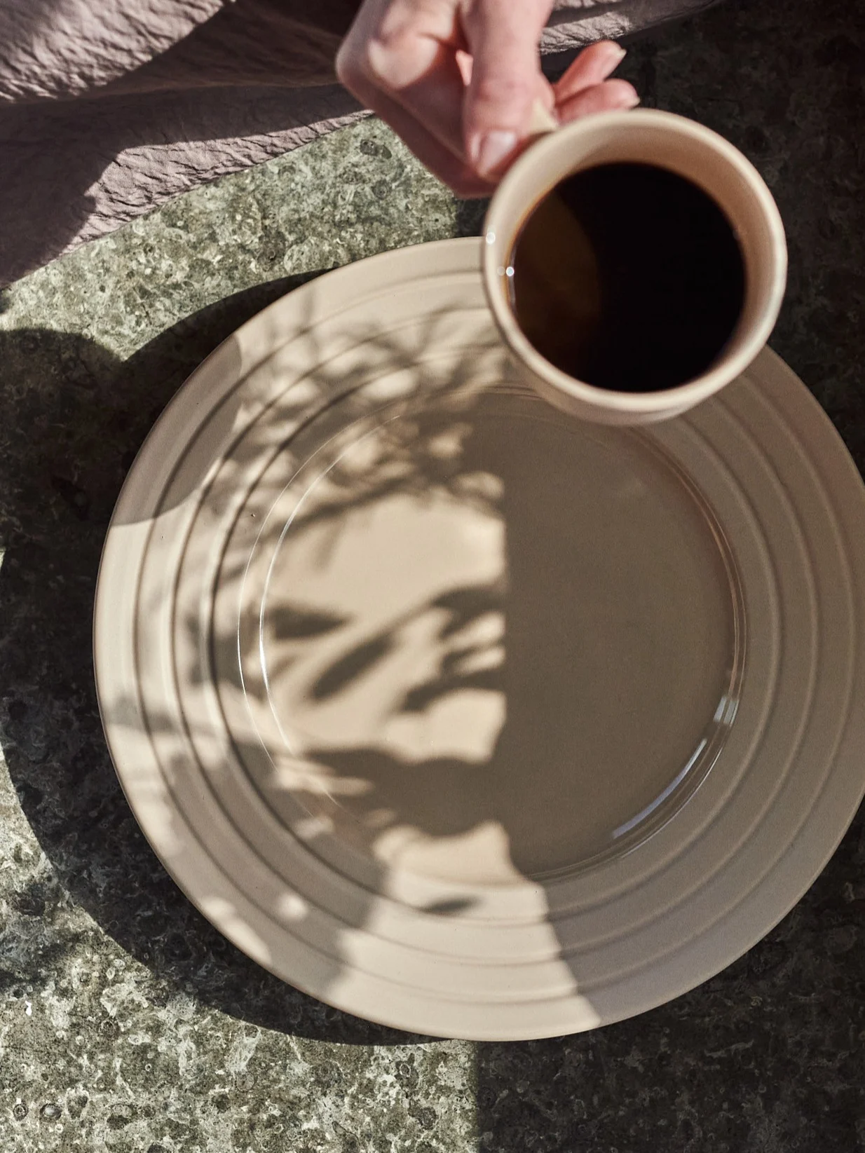 Hand holding coffee cup above beige plate with leaf shadows on a speckled table.