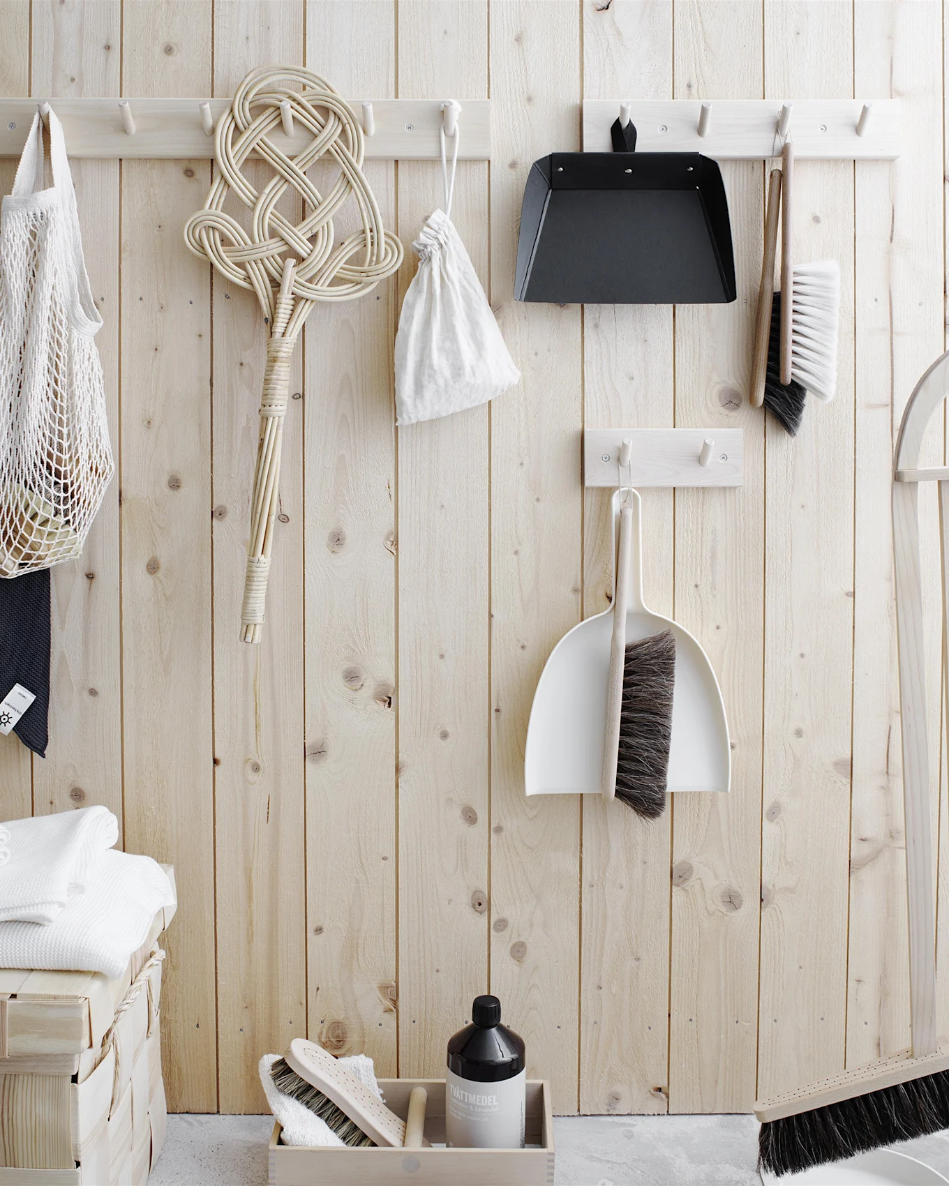 Cleaning tools and laundry essentials organized on a light wood wall. Includes brushes, dustpans, a rug beater, net bag, towels, and detergent.