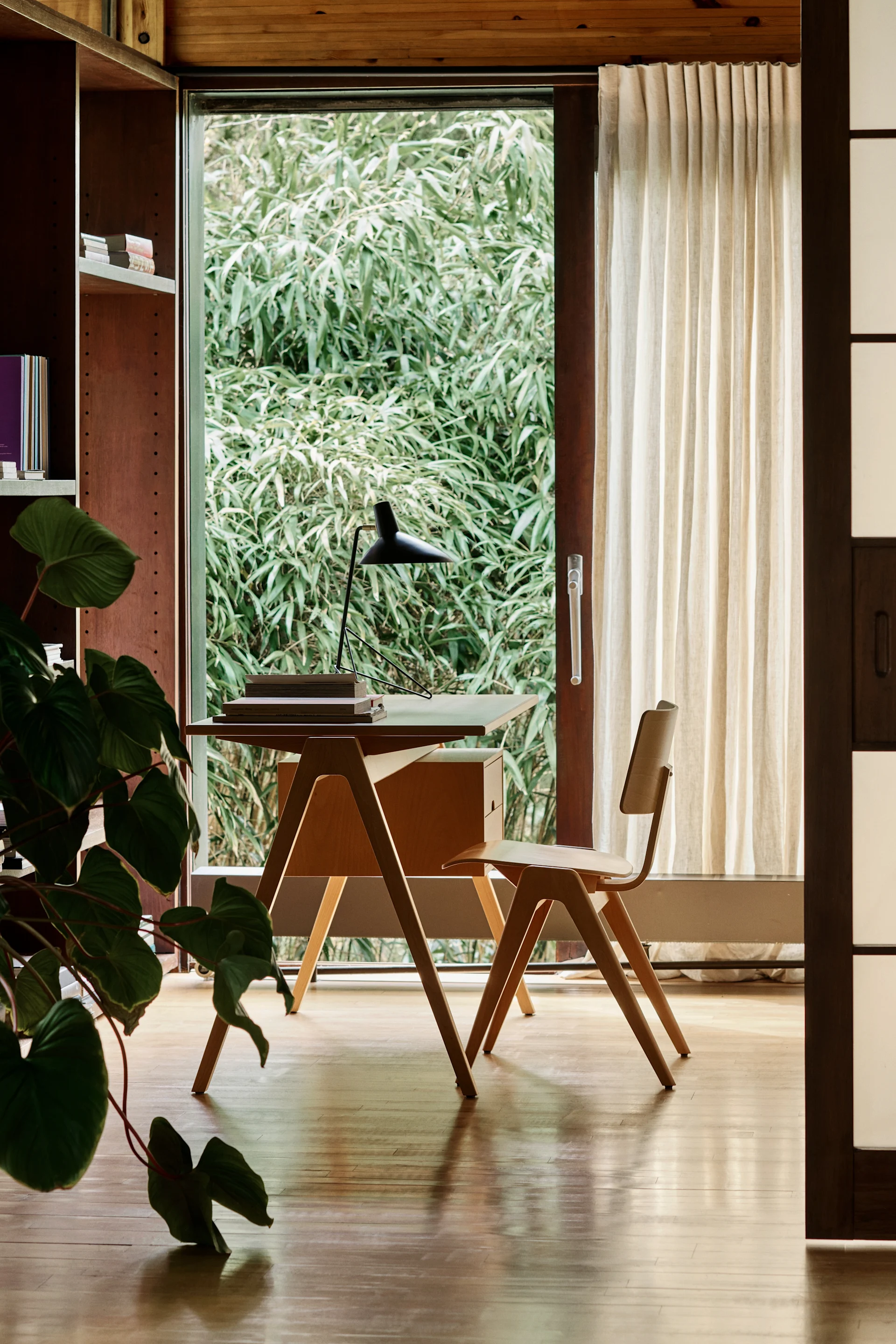 Minimalist home office with a wooden desk, chair, and lamp, overlooking green outdoor foliage.