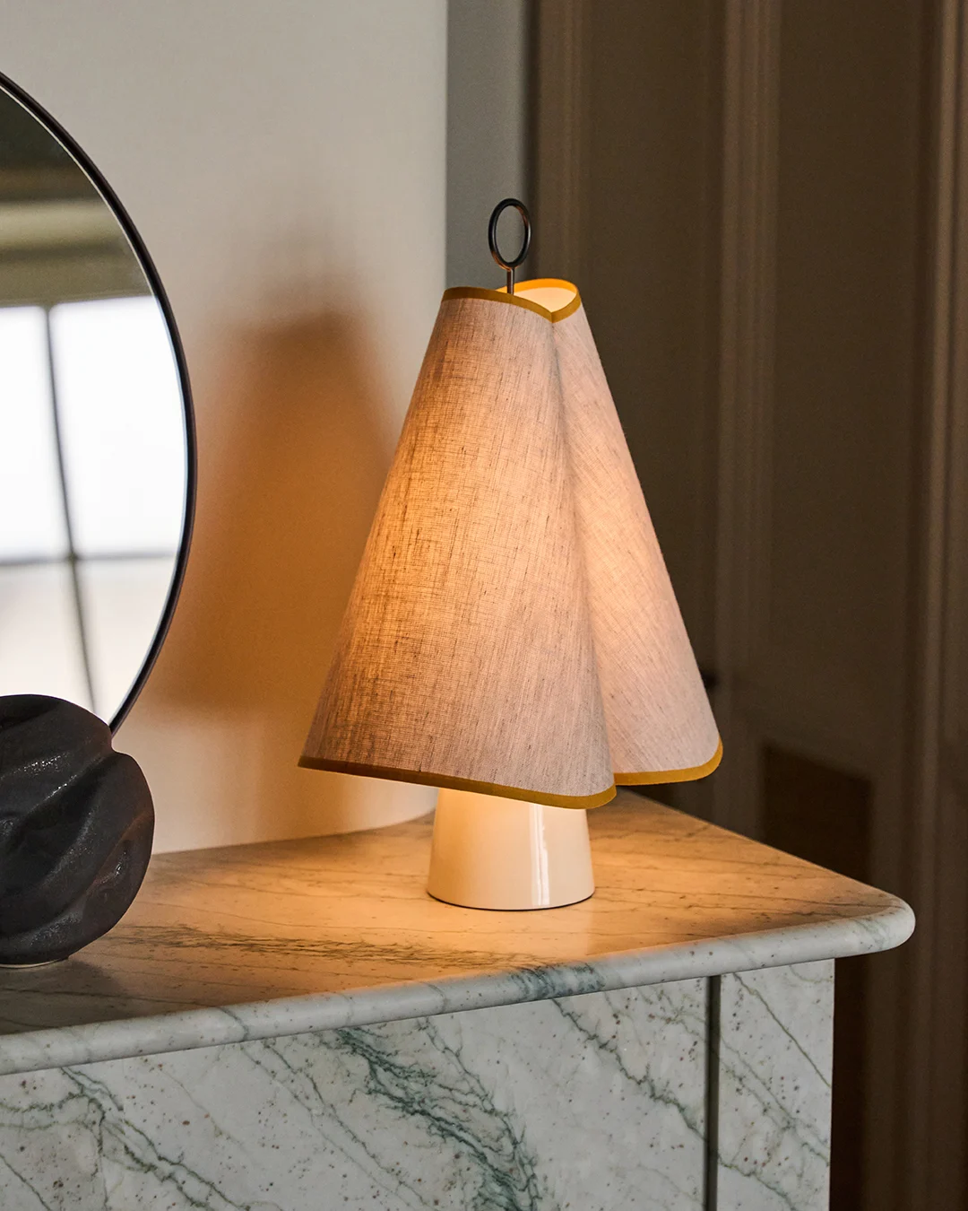 A unique table lamp with a double conical fabric shade trimmed in yellow, casting a warm glow on a green marble top.