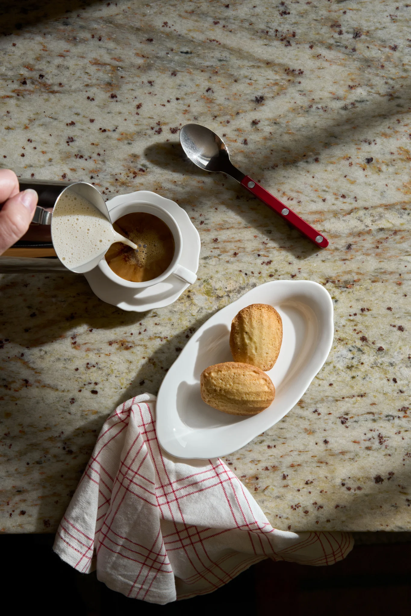 Hand pouring frothed milk into coffee. Two madeleine cookies on a white plate and a red-handled spoon rest on a granite counter.