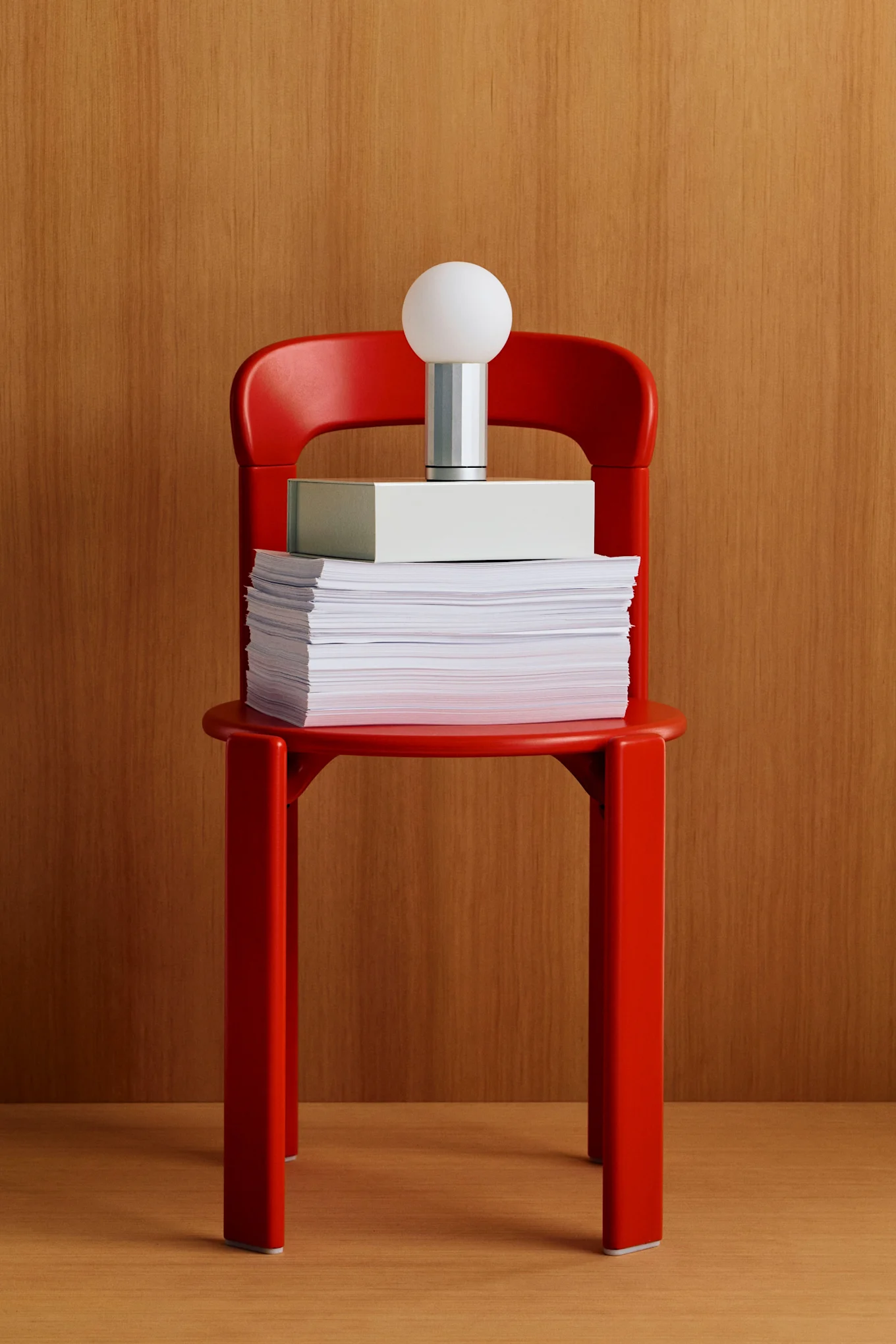 Red chair holding a stack of papers, a light gray box, and a modern lamp with a white spherical shade against a wooden background.