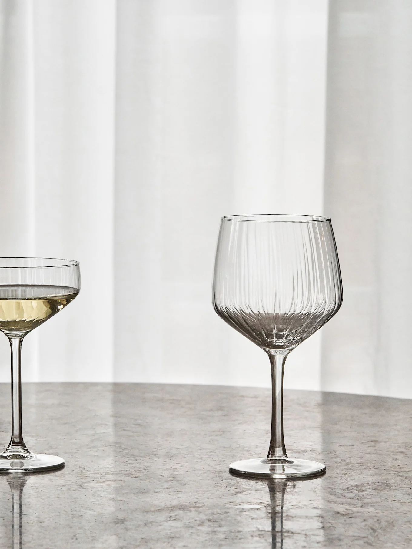 A ribbed wine glass and a coupe glass with white wine on a grey table.