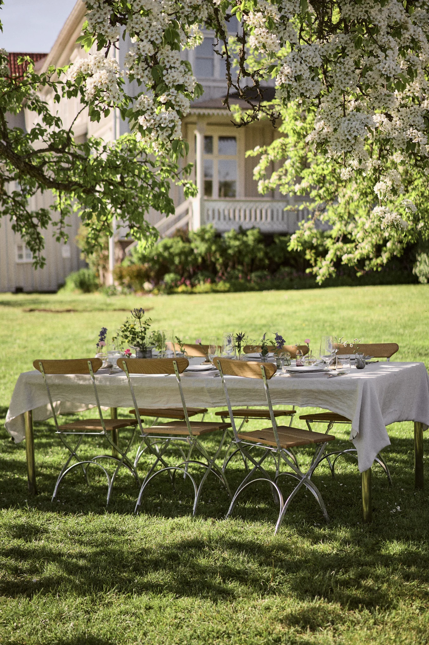 Add outdoor dining in the sunshine to your summer bucket list ideas. Here you see s table under a tree laid with a white tablecloth and a set of Grythyttan chairs.  
