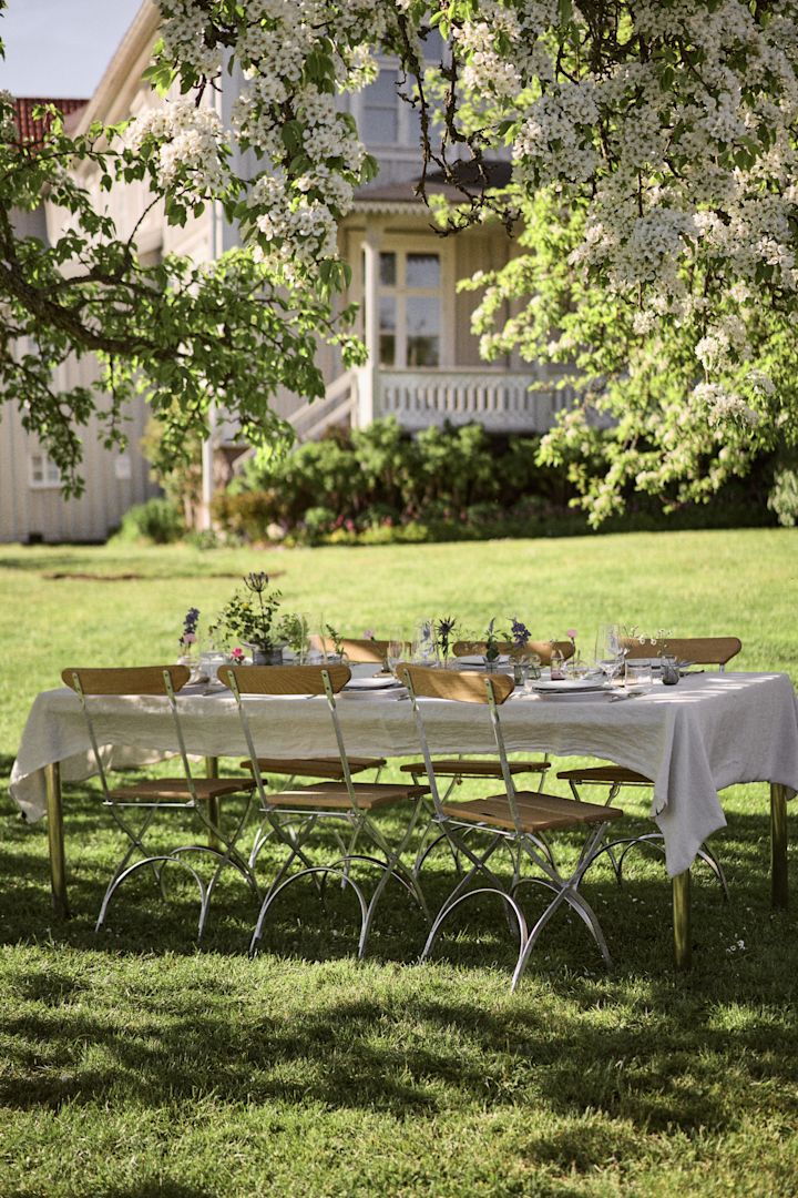 Add outdoor dining in the sunshine to your summer bucket list ideas. Here you see s table under a tree laid with a white tablecloth and a set of Grythyttan chairs.  