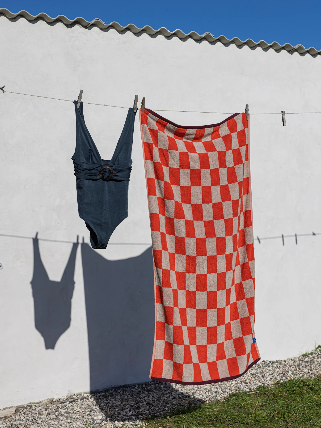 A dark blue one-piece swimsuit and a red and beige checkered beach towel hang on a clothesline against a white wall.